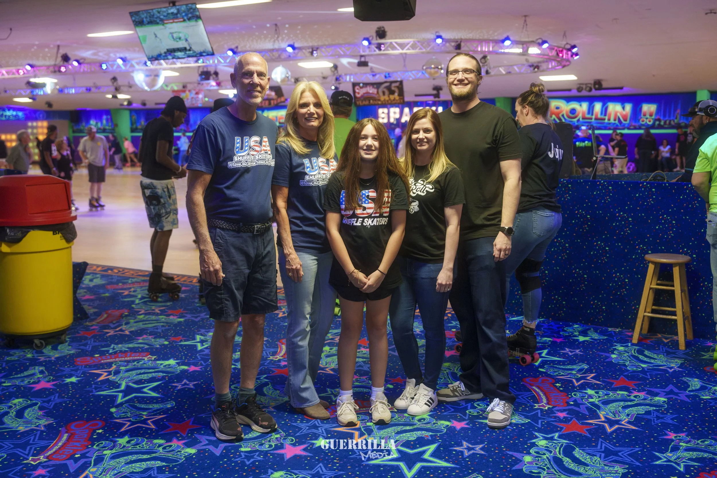 Group of six people posing inside roller skating rink, colorful neon lights, and retro decor in background.