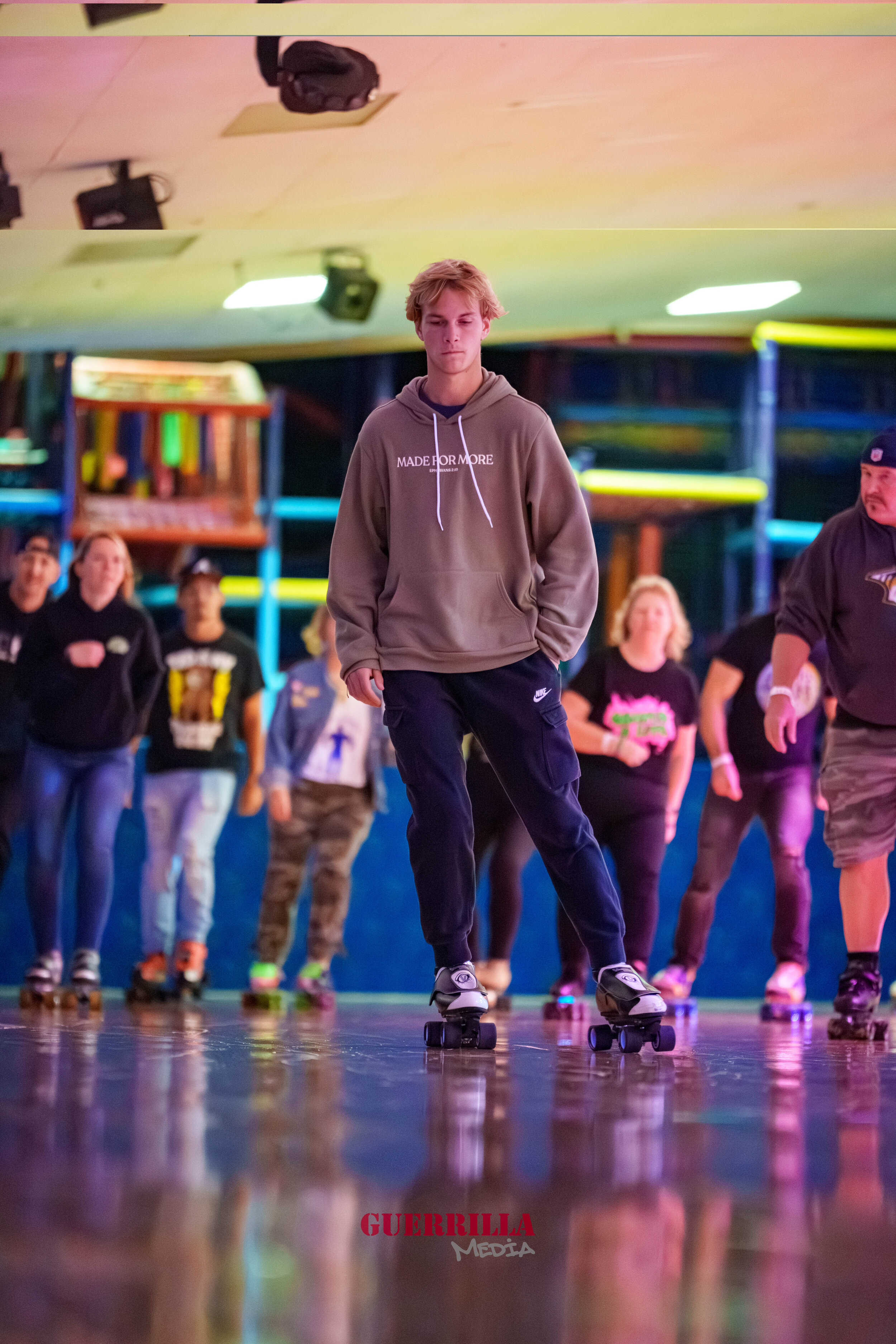 Young man roller skating at an indoor roller rink, with other skaters in the background and colorful arcade or playground structures. The young man is wearing a beige hoodie and black pants.
