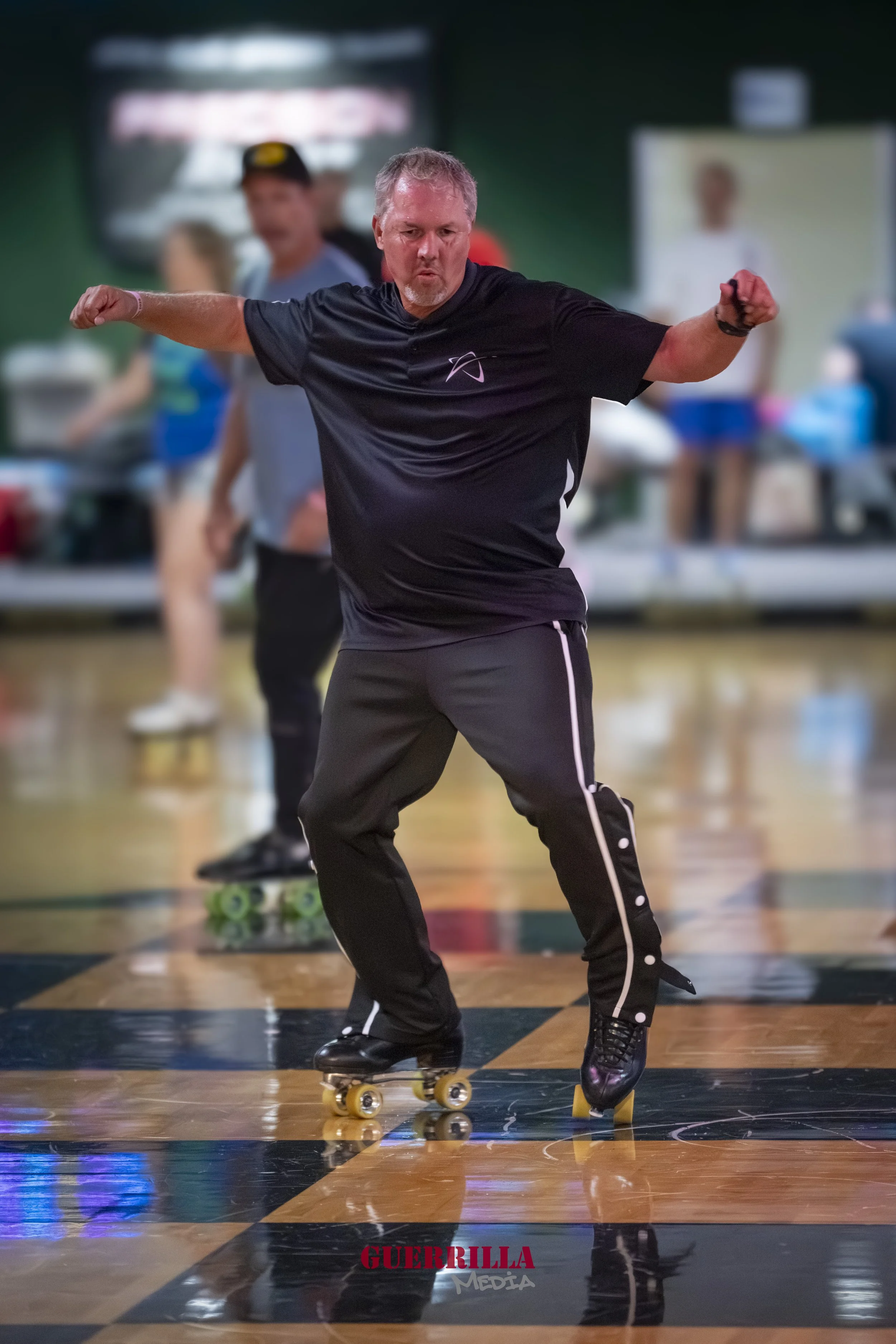 A man roller skating indoors on a polished wooden floor, with his arms extended and a focused expression, while other people are blurred in the background.