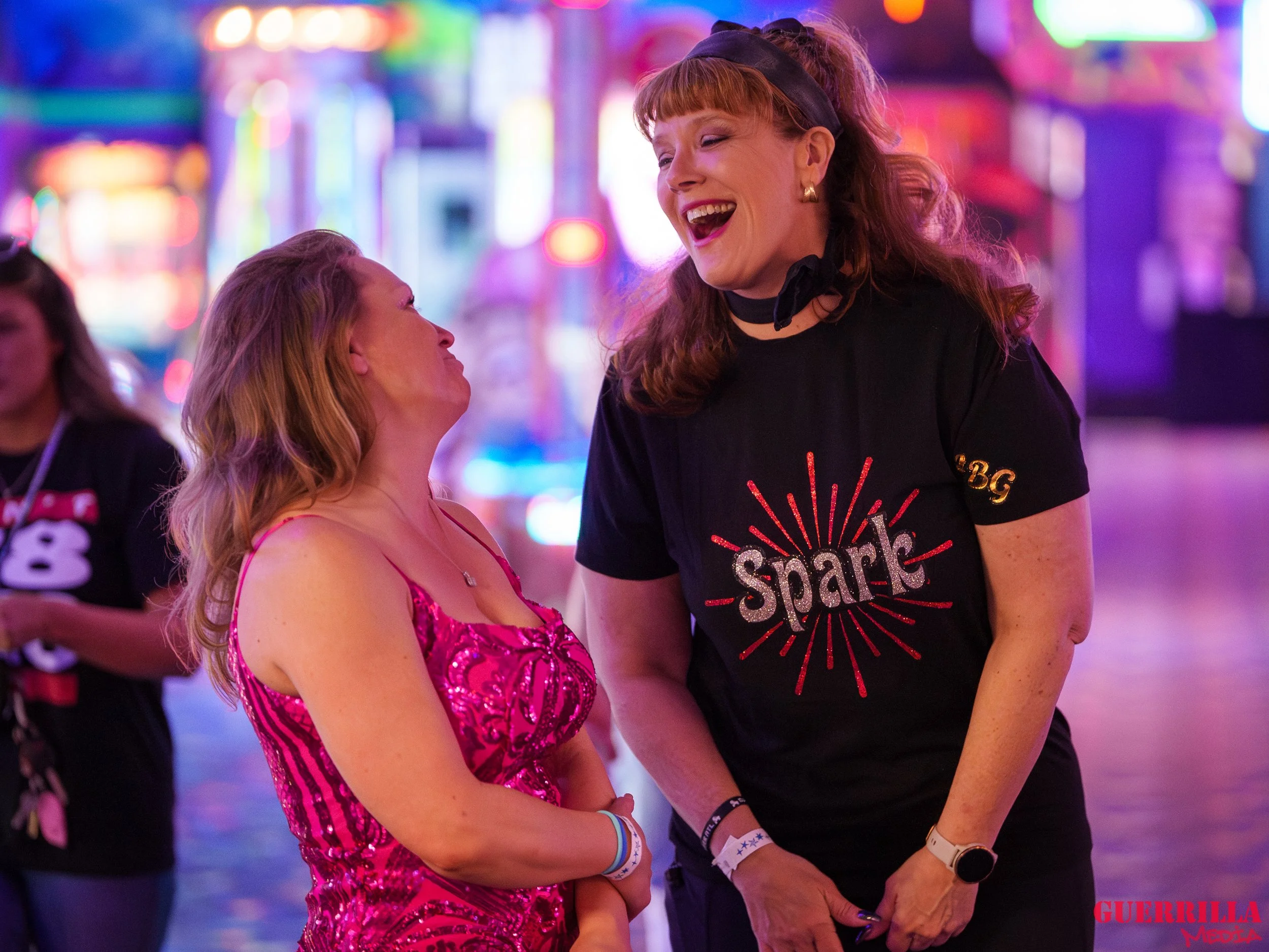 Two women, one in a pink dress and one in a black t-shirt with 'Spark' on it, laughing and smiling at each other in a brightly lit, colorful, outdoor nightlife setting.