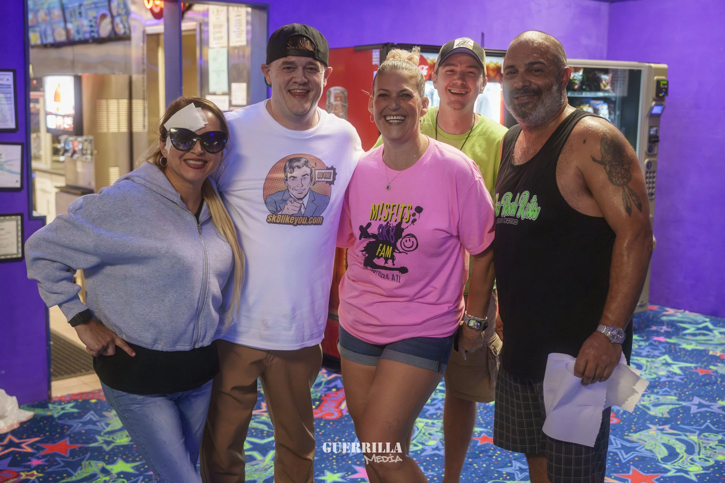 Group of five people smiling and posing together inside a colorful arcade or entertainment venue with purple walls and neon lights.