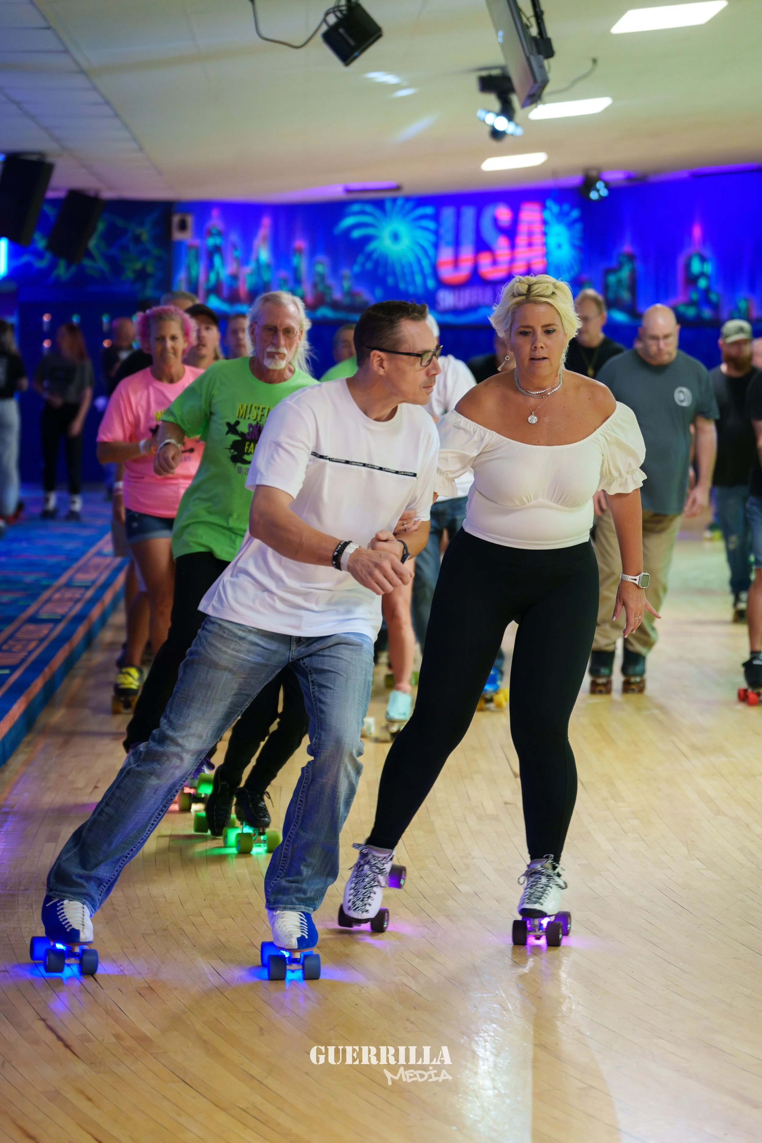 People roller skating in an indoor roller rink decorated with USA-themed neon signs and fireworks on the wall.