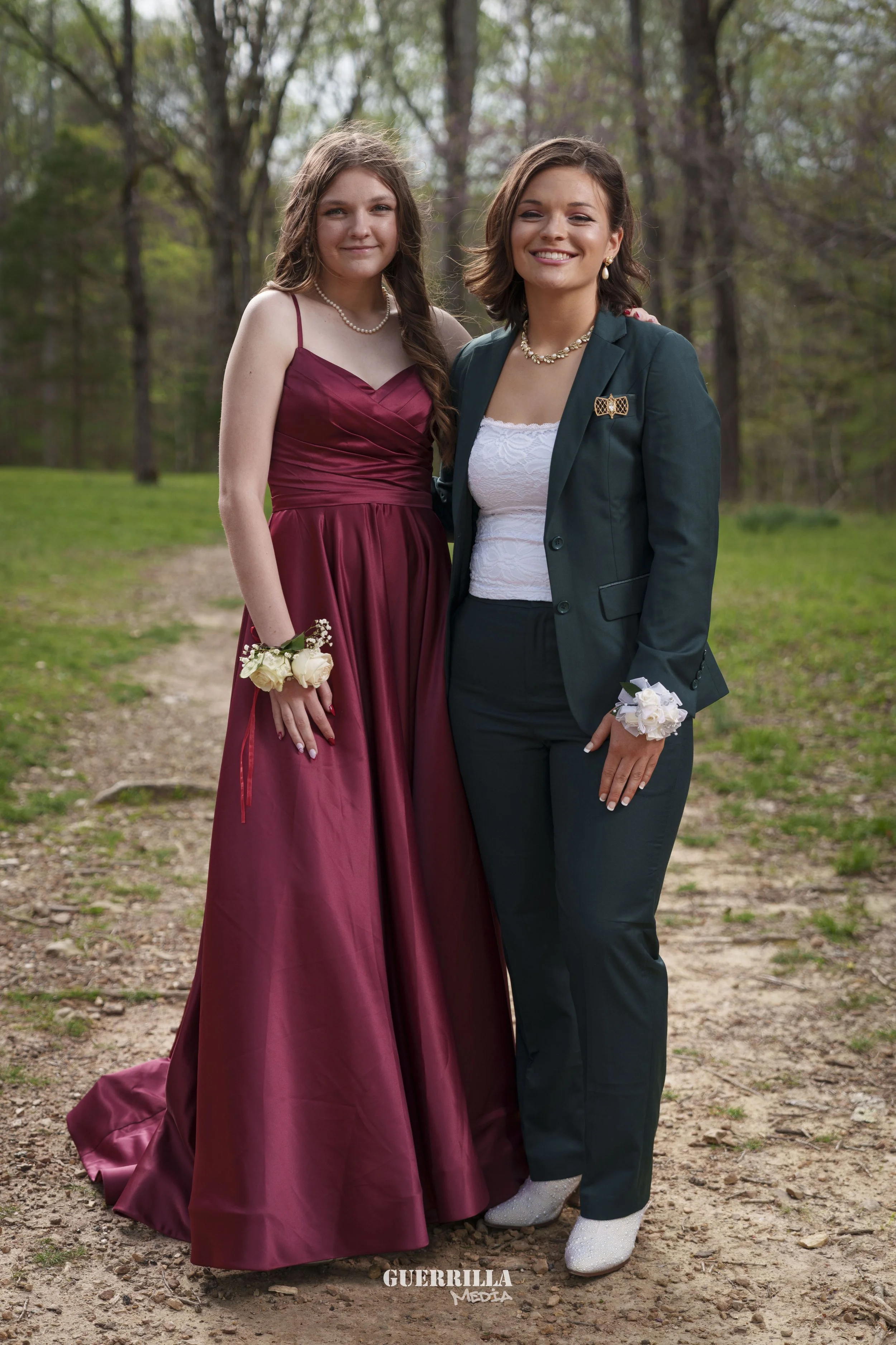 Two women dressed in formal attire standing outdoors on a dirt path in a wooded area. One wears a long, burgundy gown with a corsage, and the other wears a dark green suit with a white lace top and a corsage. Both are smiling.
