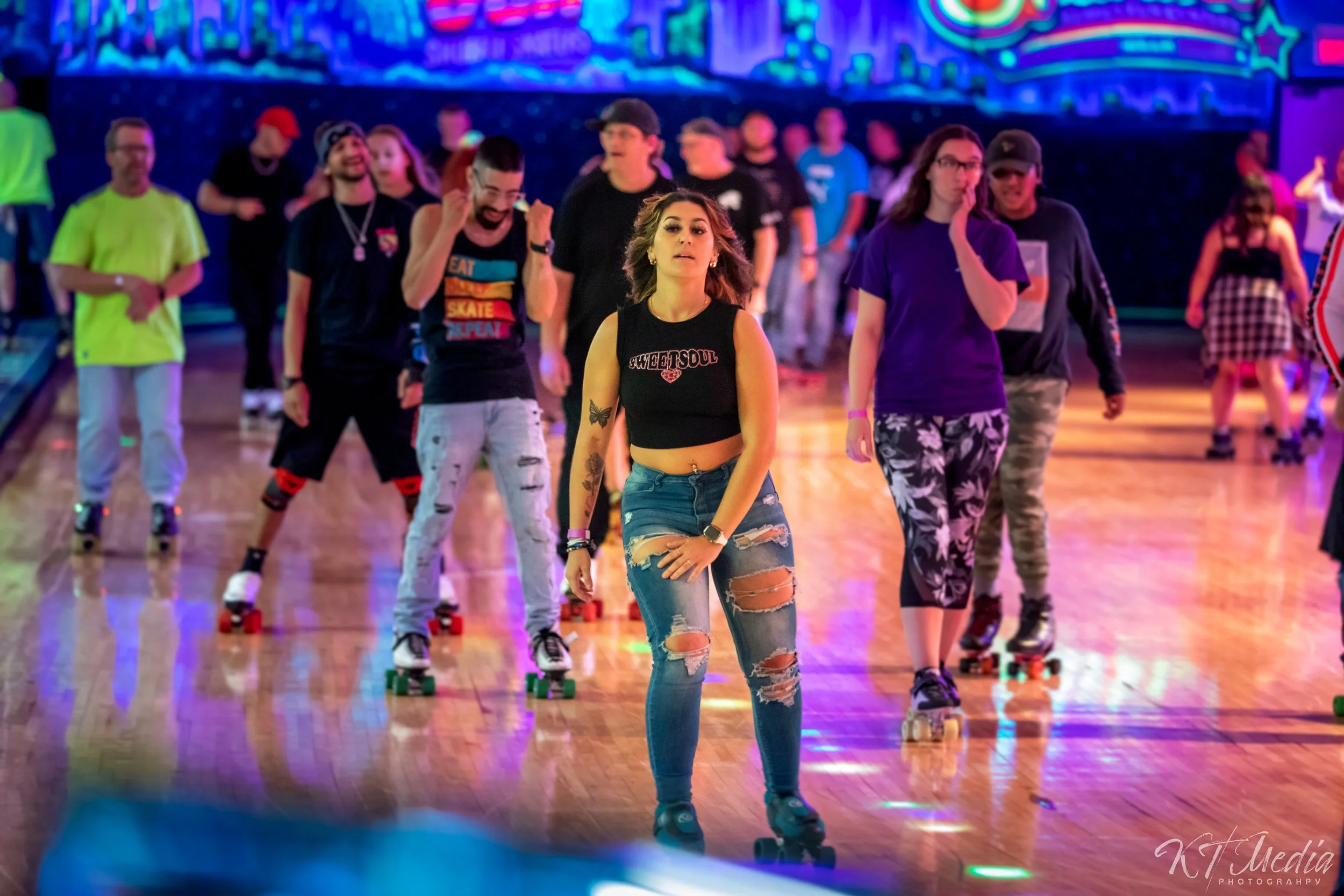 People roller skating in a roller rink, including a young woman with torn jeans and a black crop top at the front, and others behind her on roller skates, with colorful lights and a digital display in the background.