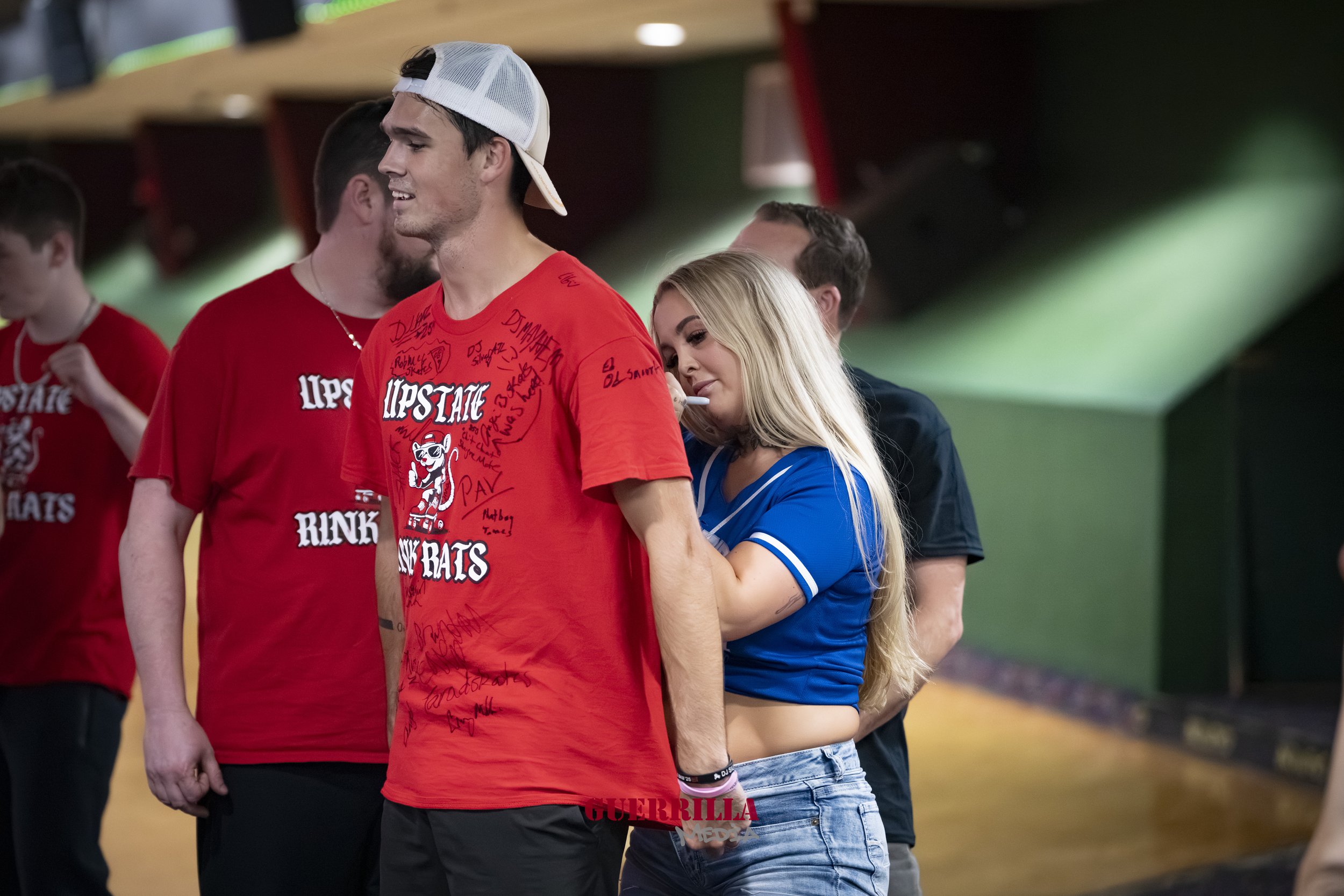 A group of young adults at an indoor roller skating rink. A young man in a red T-shirt and white baseball cap is standing with his eyes closed and a slight smile. A blonde woman in a blue top is behind him, leaning on his back with her eyes closed an