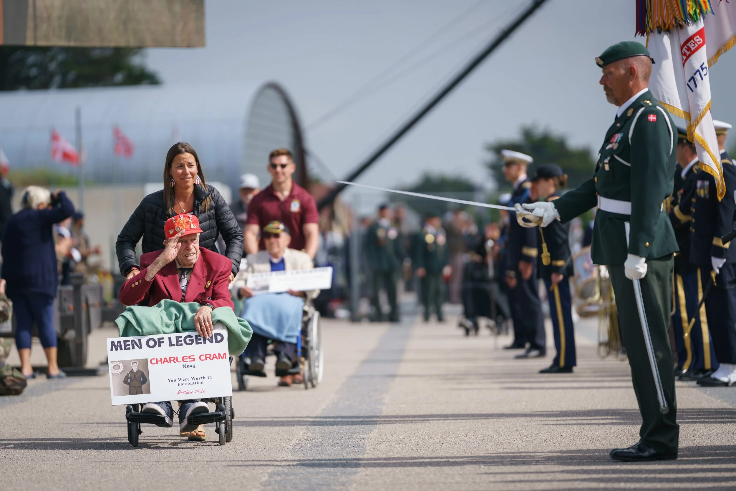 A woman in a wheelchair salutes while an elderly man in a wheelchair salutes back; they are at a military ceremony with a marching band, and a soldier in uniform salutes them.