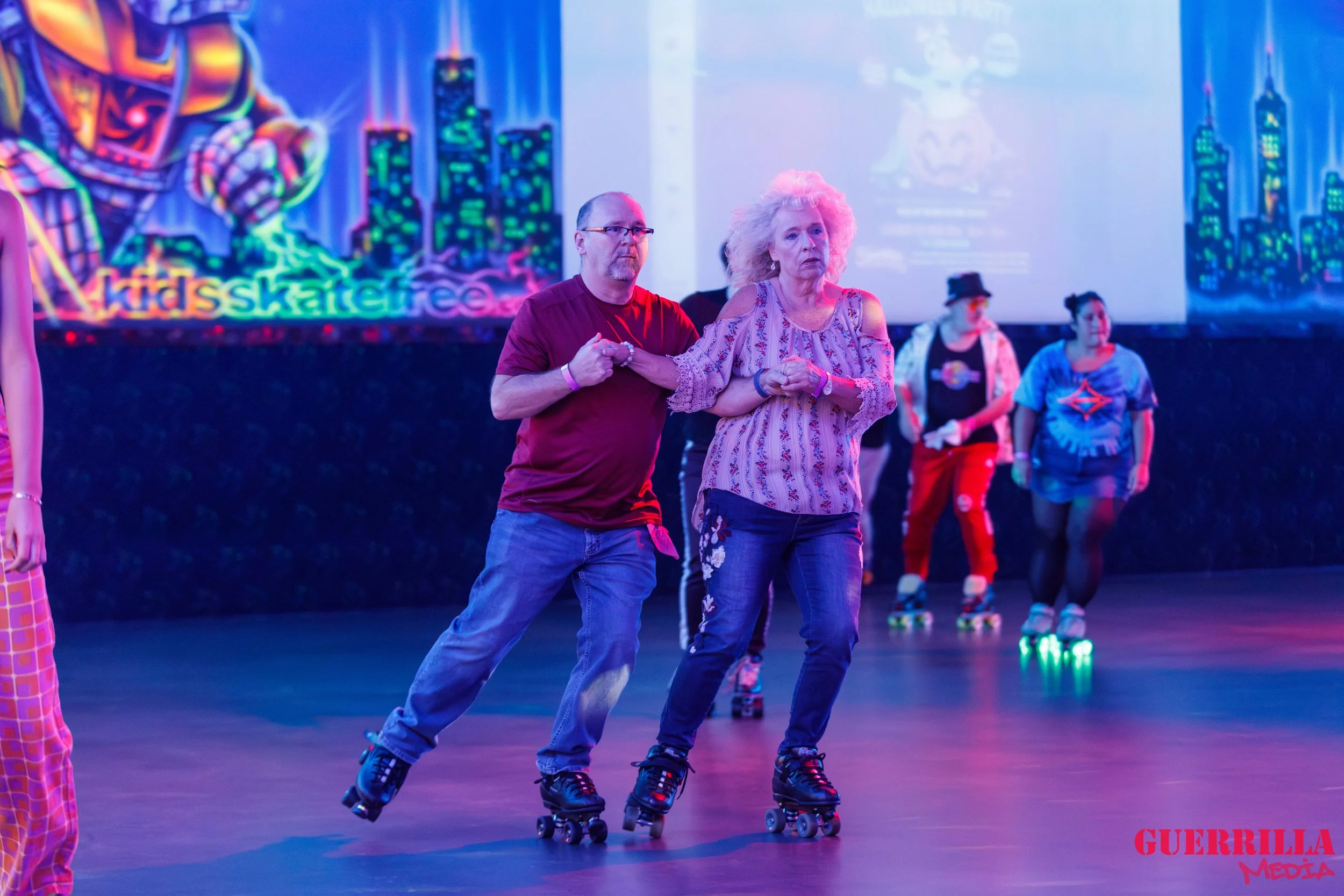 People roller skating in indoor rink with neon cityscape murals. One man helping a woman skate.