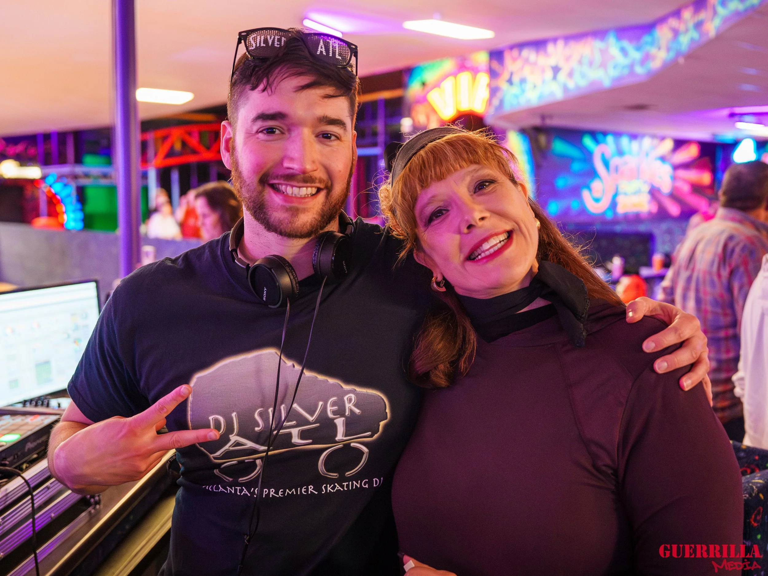 A young man wearing headphones around his neck and sunglasses on his head making a peace sign with his right hand, and a woman with red hair and a black shirt smiling and leaning into him, in an indoor neon-lit arcade or entertainment venue.