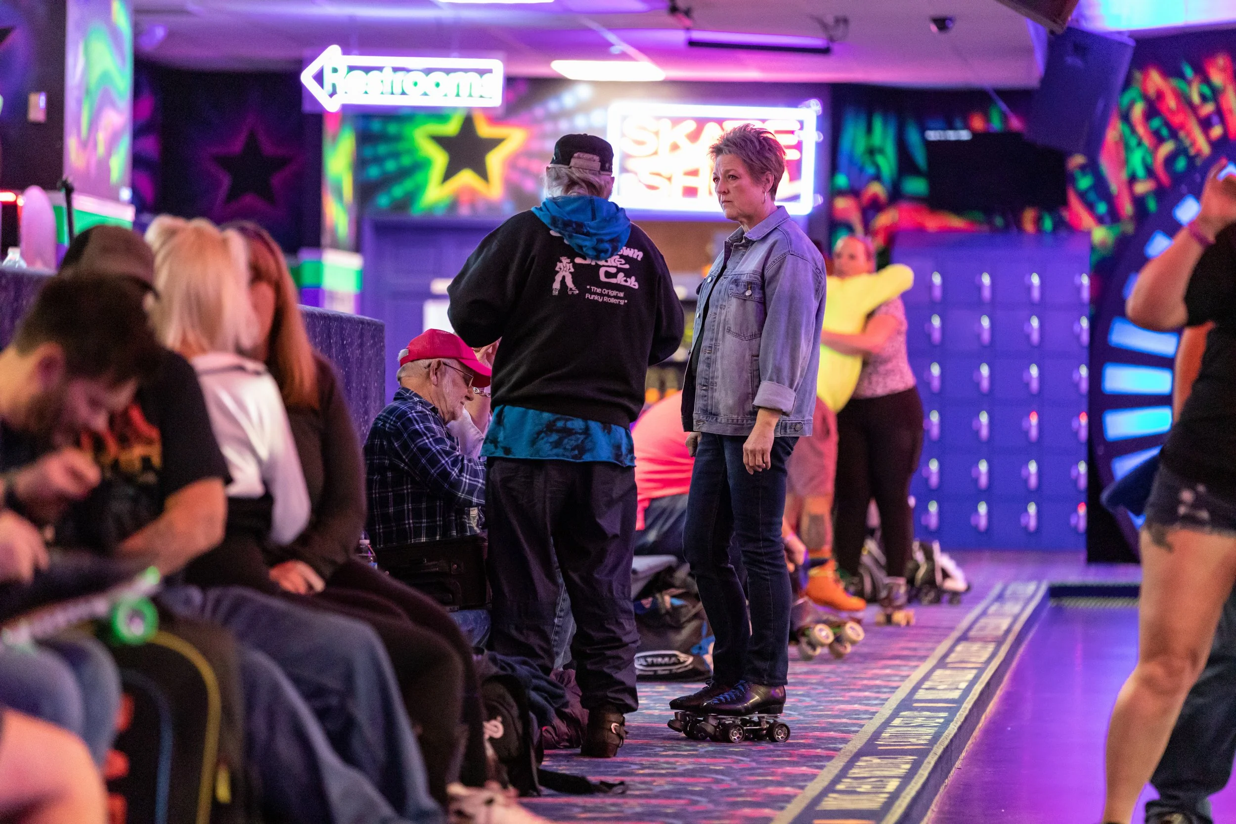 People sitting and standing at an indoor roller skating rink with colorful neon signs and vibrant lighting.