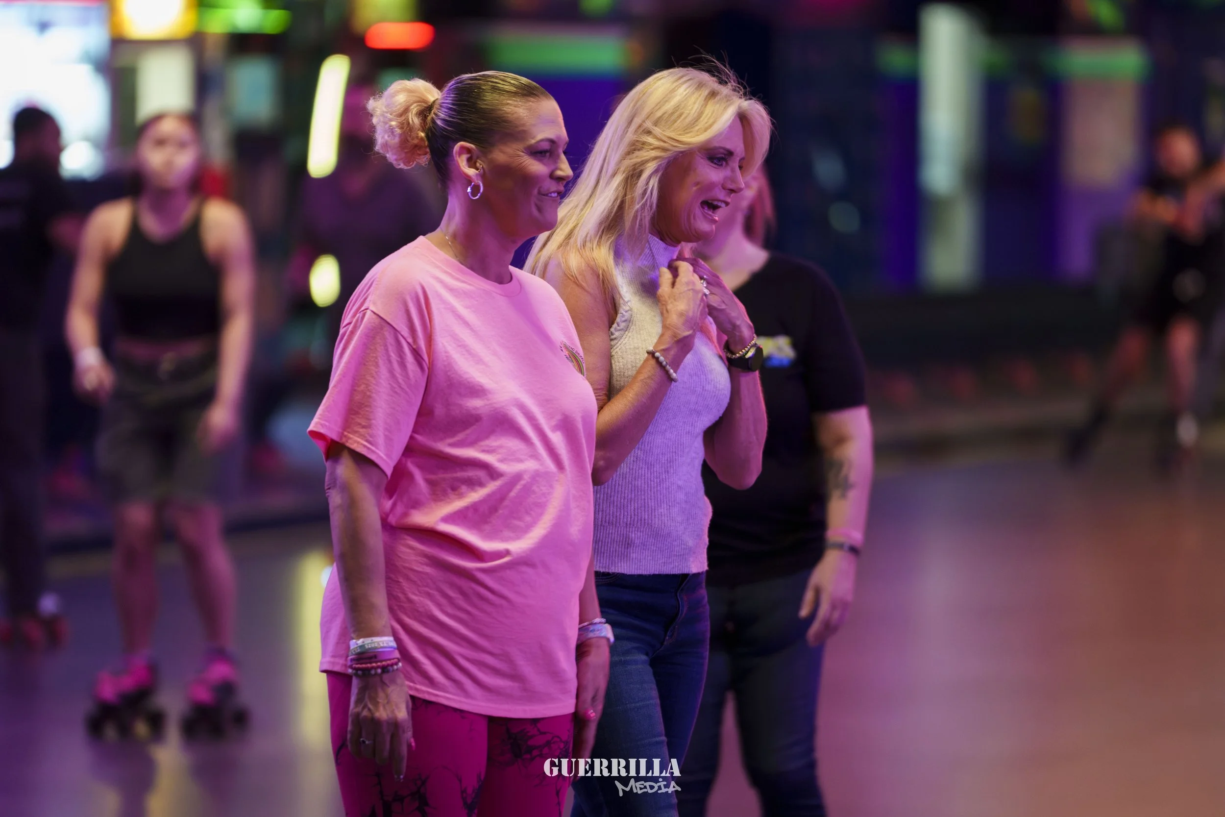 Two women, one in a pink shirt and pants and the other in a gray sleeveless top, are standing in a roller skating rink with neon lights, looking happy and emotional, surrounded by other people roller skating.