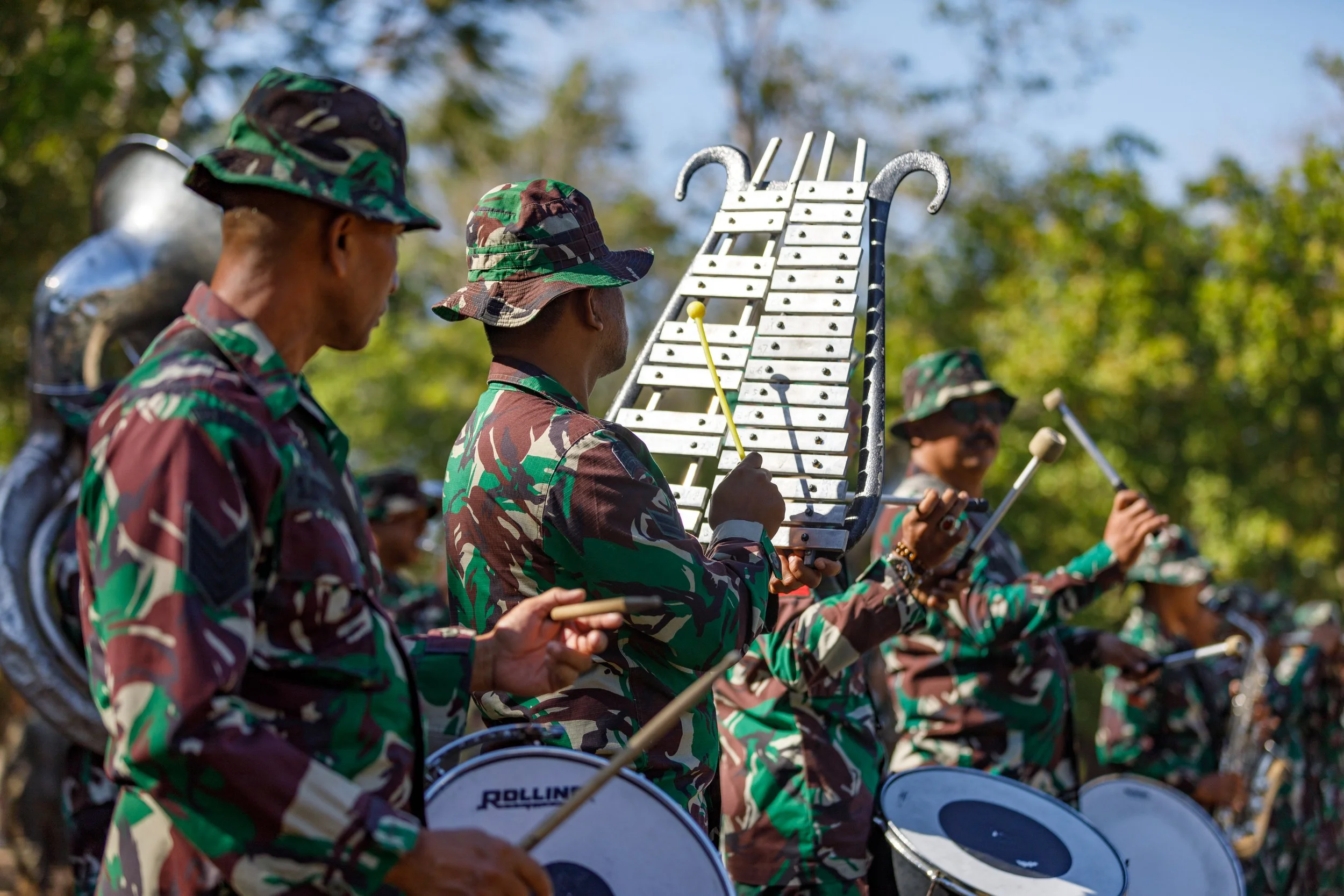 Military band wearing camouflage uniforms playing percussion and wind instruments outdoors, with trees in the background.