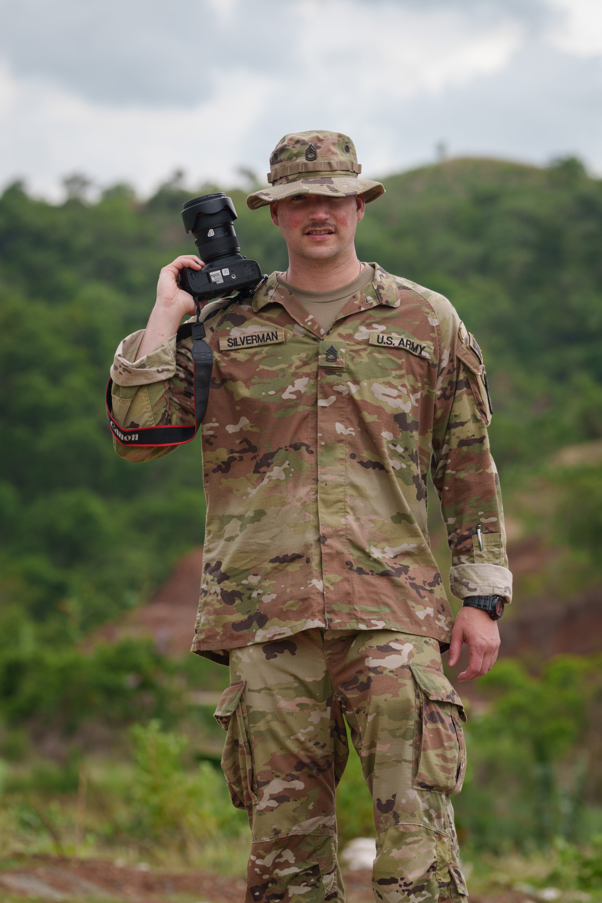 A U.S. Army soldier in camouflage uniform holding a camera with a telephoto lens over his shoulder, standing outdoors with green trees and hills in the background.
