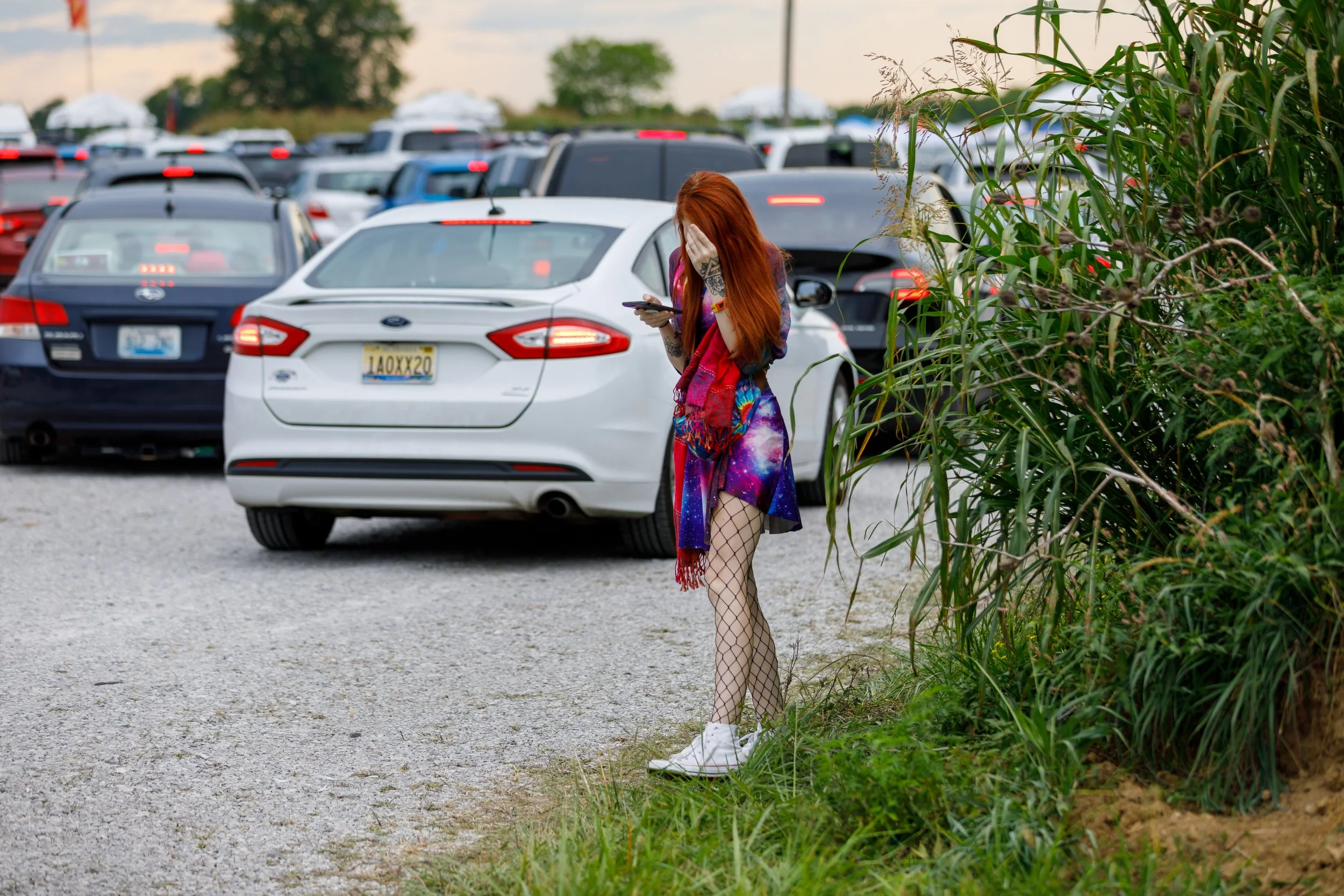 A young woman with red hair, dressed in a colorful galaxy-themed dress, fishnet stockings, and white sneakers, stands near a grassy area while looking at her phone at a crowded parking lot with many cars.