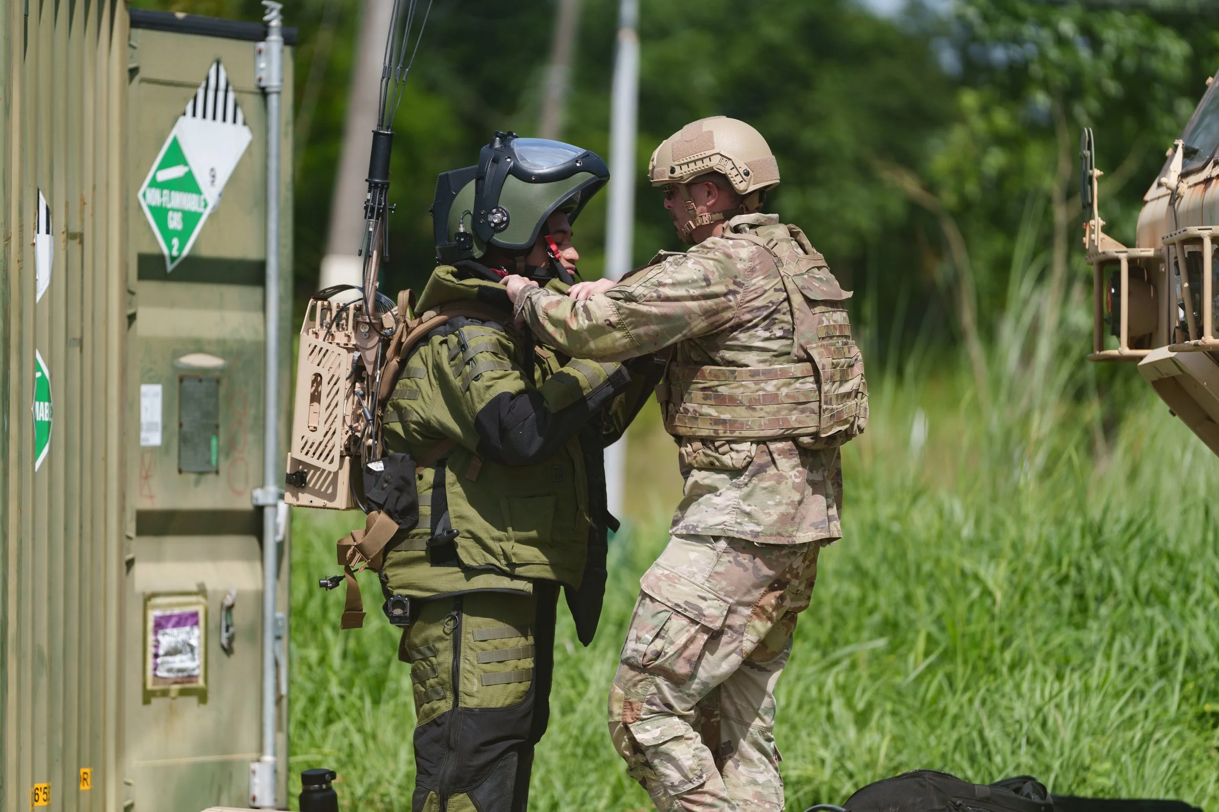 A soldier helps a pilot put on a helmet after landing, in a grassy outdoor area with military equipment nearby.