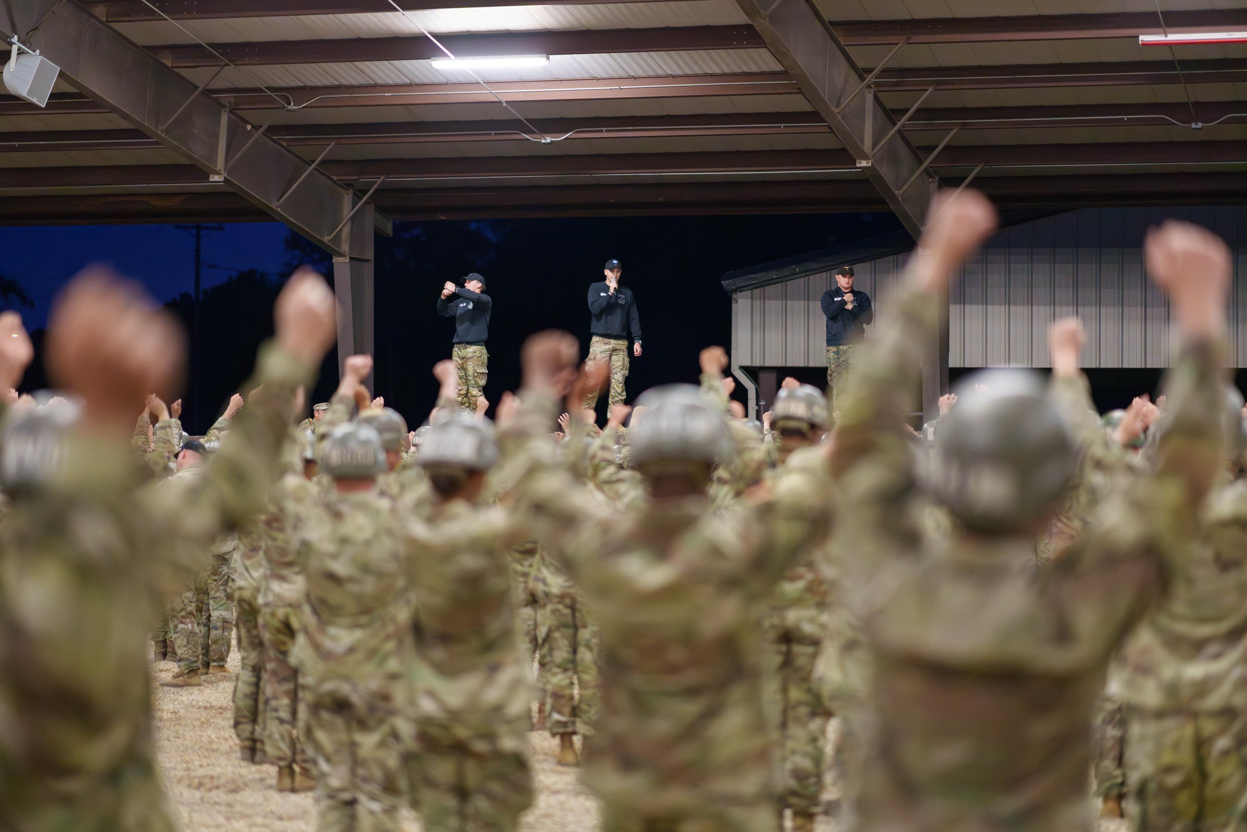 Military personnel in camouflage uniforms standing in formation and raising their fists, watching a stage where three officers are speaking into microphones inside a large covered structure.