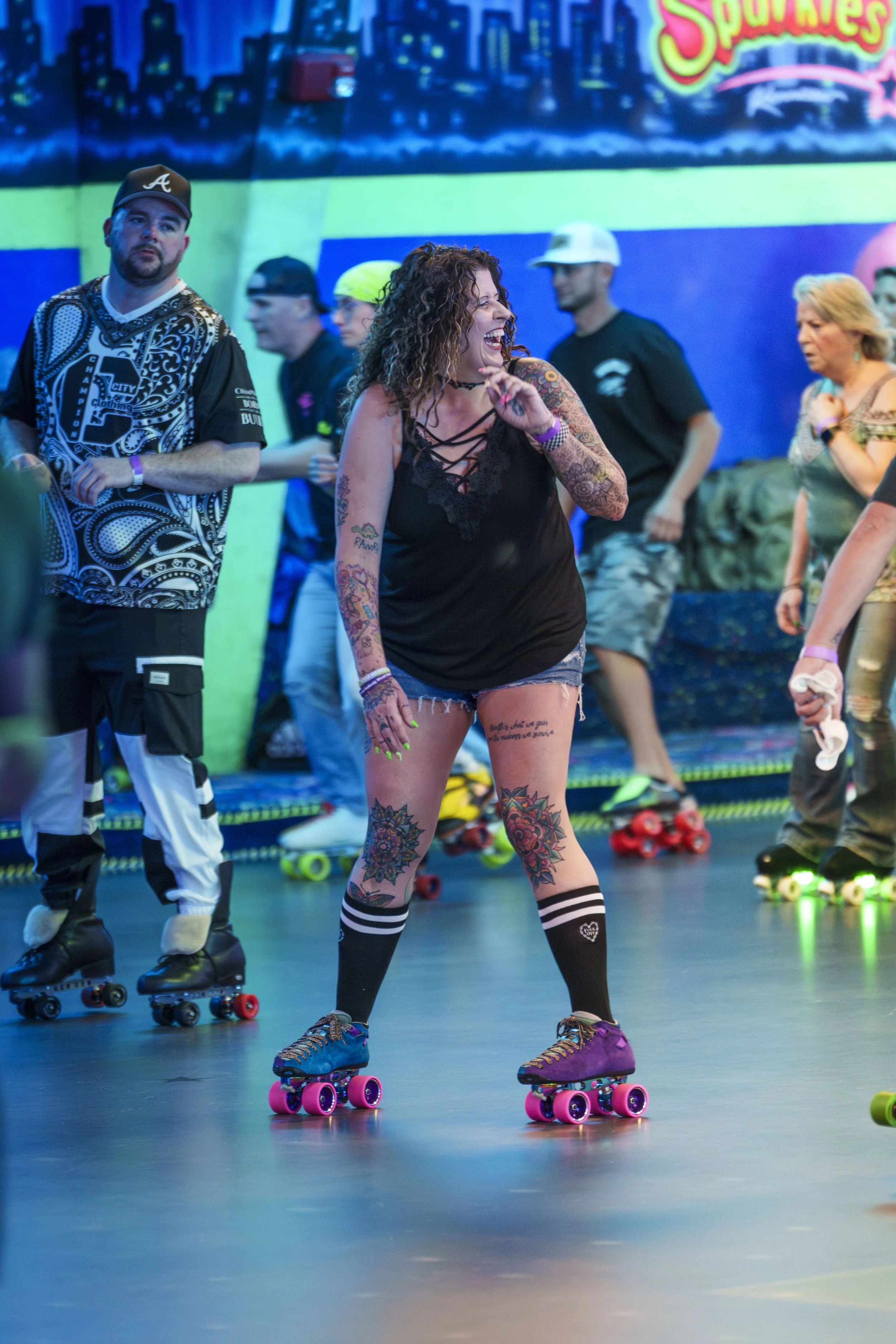 People roller skating at an indoor skating rink, with a woman smiling and laughing in the foreground, wearing a black top, shorts, and striped knee-high socks, tattoos visible on her legs and arms.