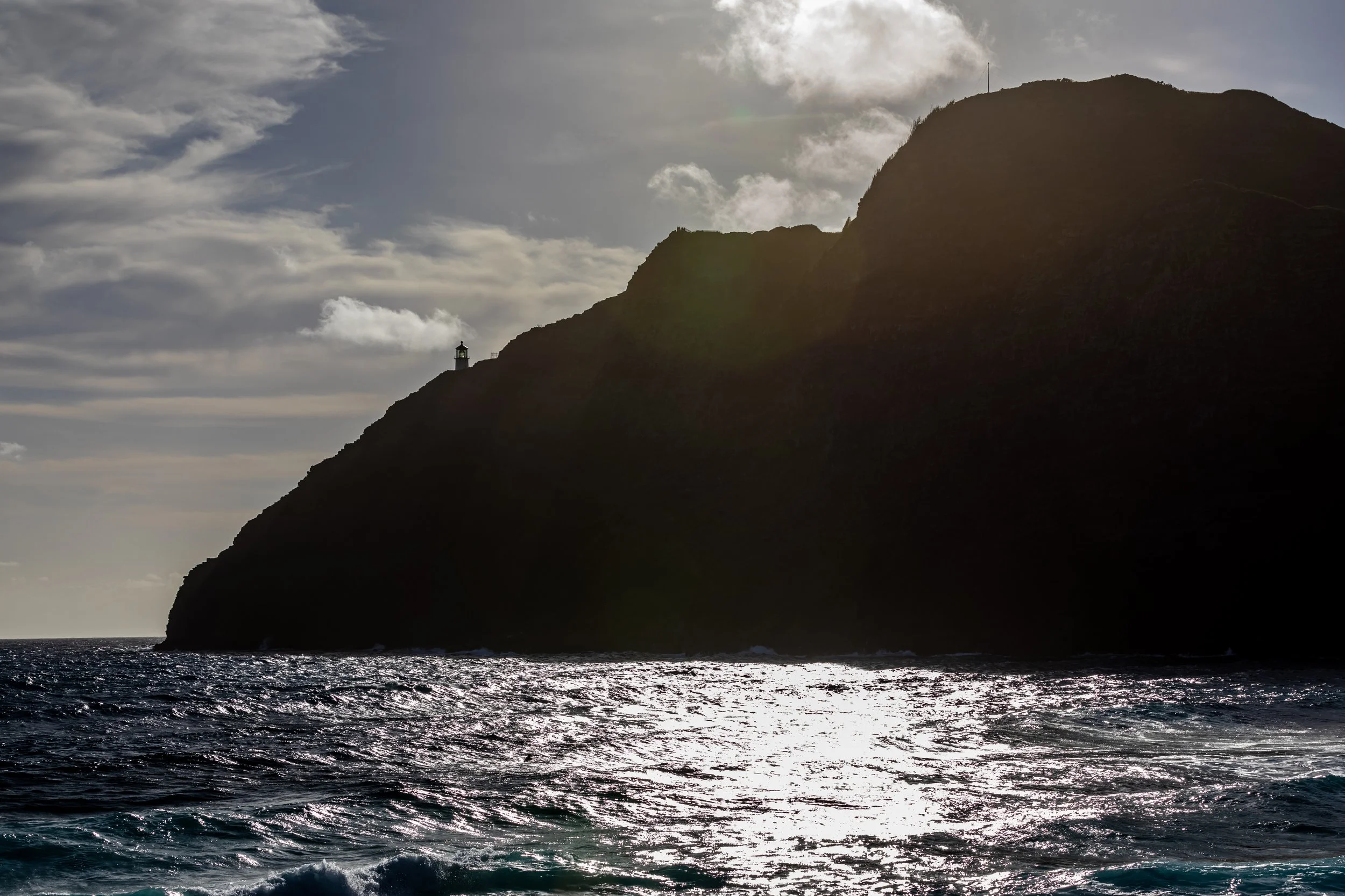 Sunset over a rocky coastal hillside with a lighthouse silhouette on top of the cliff, reflecting on the ocean waves.