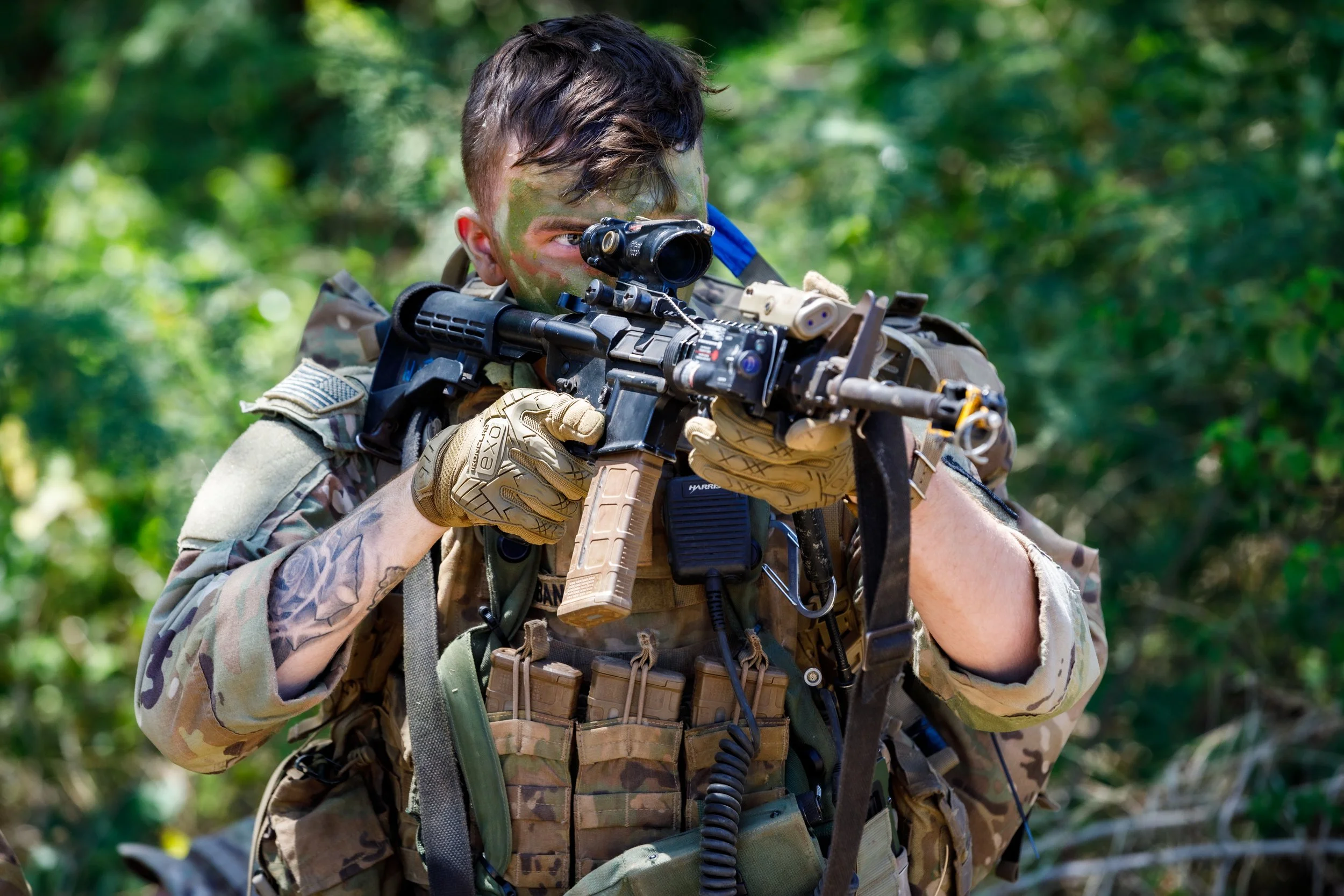 Soldier in camouflage uniform aiming a rifle with a scope in a wooded area.