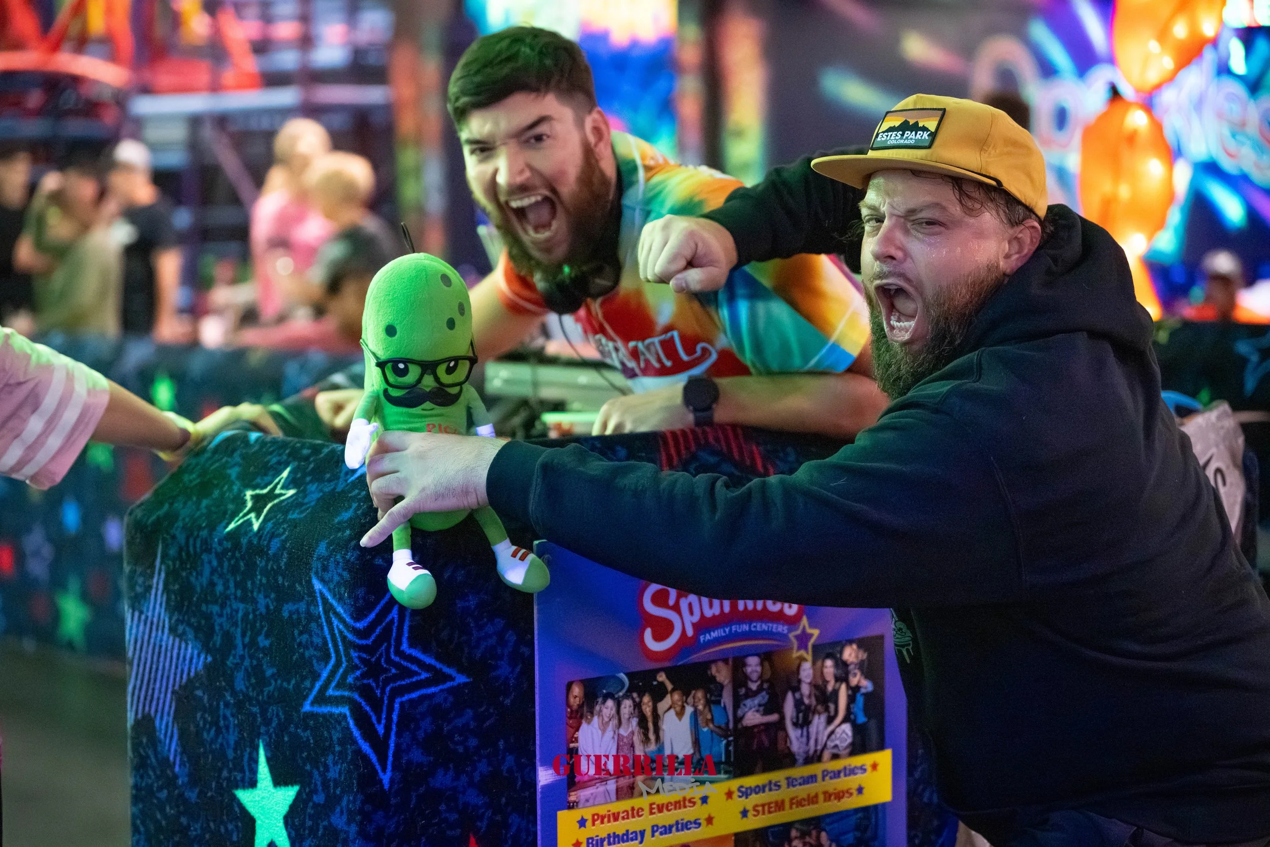 Two men with beards celebrating at an amusement park or arcade, one wearing a rainbow tie-dye shirt and the other in a black hoodie and yellow cap, posing with a green plush toy of a pickle character wearing glasses, a mustache, and sneakers, in fron