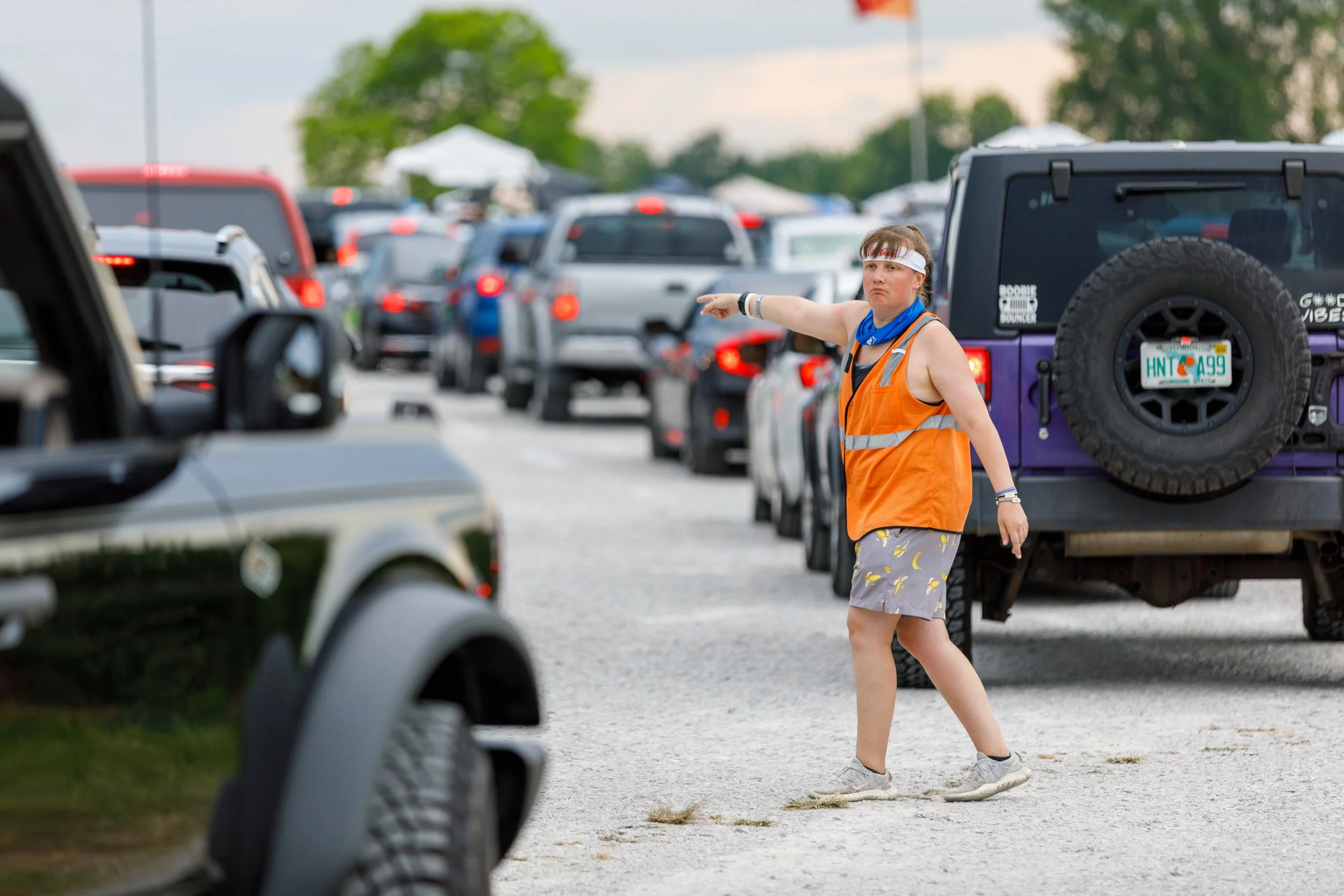 A crowd of cars parked in a lot with a woman in an orange safety vest, shorts with a banana pattern, white sneakers, and a headband, pointing to the left.