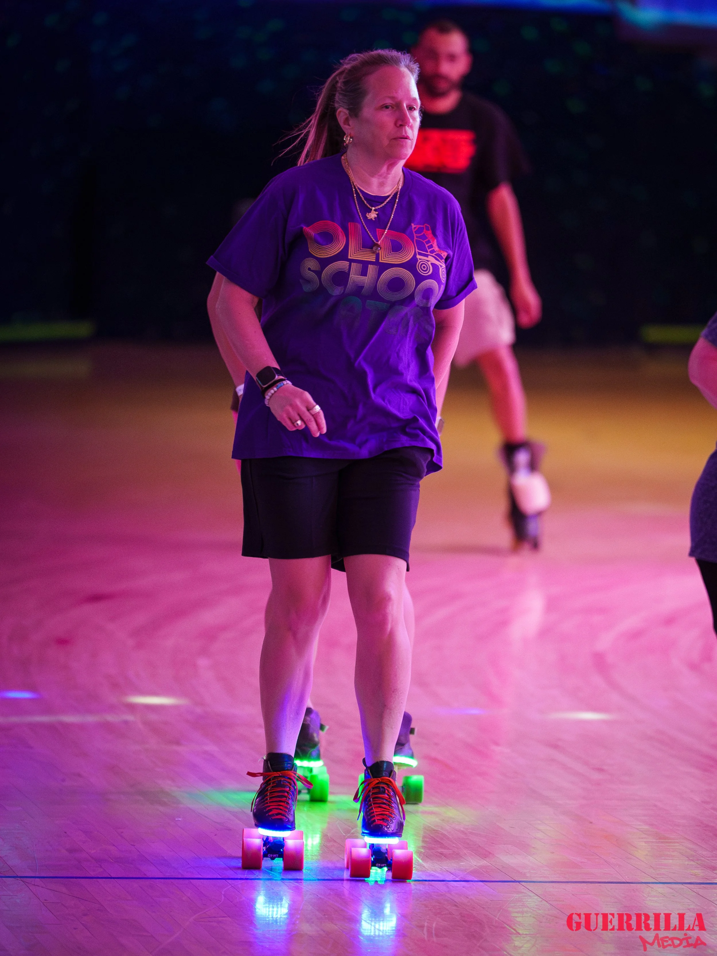 Woman skating on roller skates with colorful lights in a dark indoor rink, wearing a purple T-shirt and black shorts.