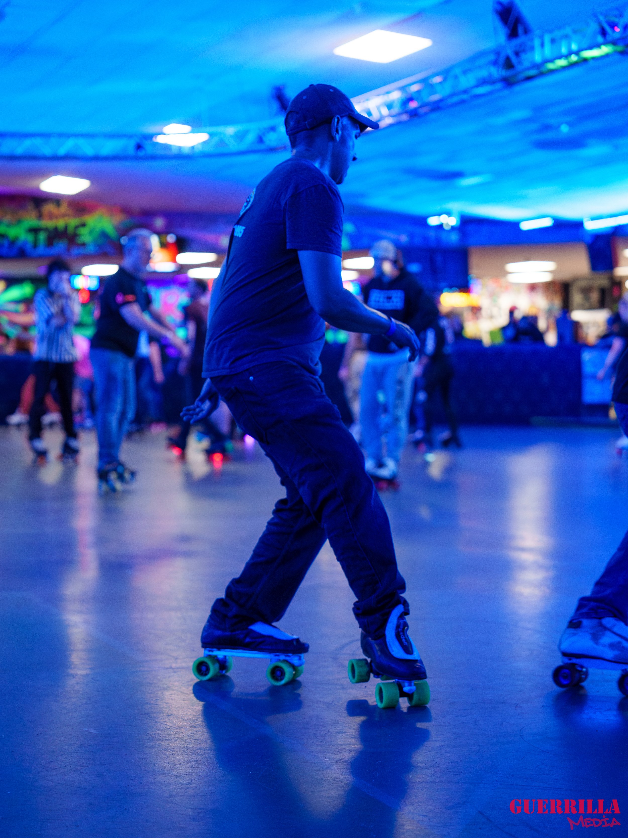 Person roller skating in an indoor rink with blue lighting, with other skaters in the background.
