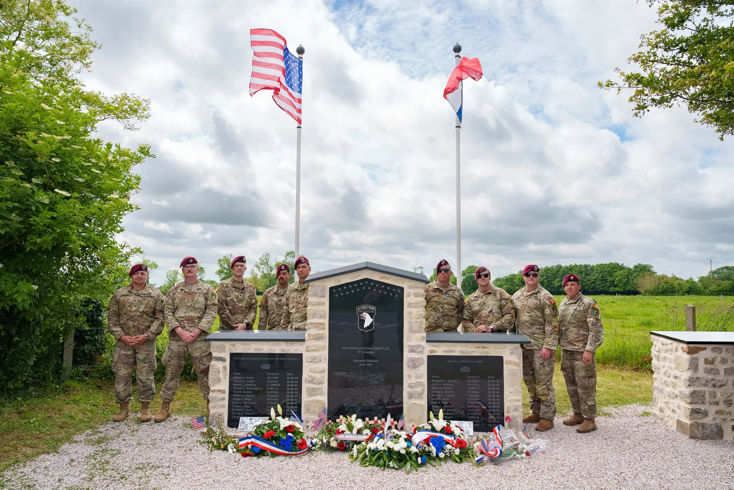 Group of soldiers standing in front of a memorial with American flags, floral wreaths, and plaques, with a cloudy sky in the background.