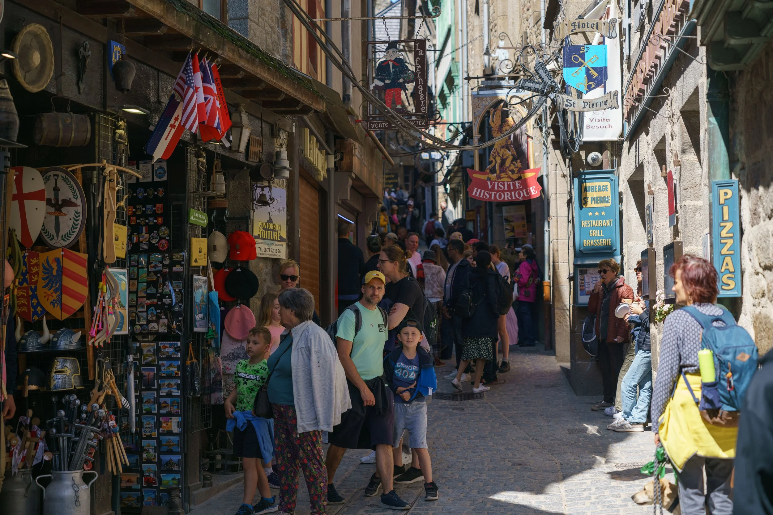 Crowded narrow street with various shop signs, people walking and shopping, souvenirs and hats on display.