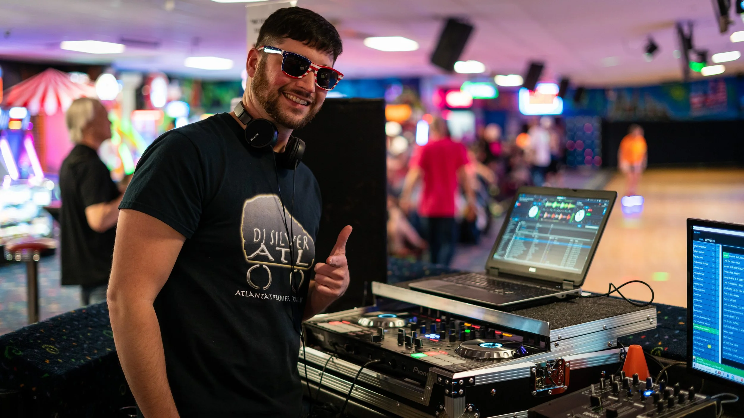 Men DJ wearing sunglasses, standing in front of DJ equipment, giving a thumbs-up at an indoor entertainment venue.