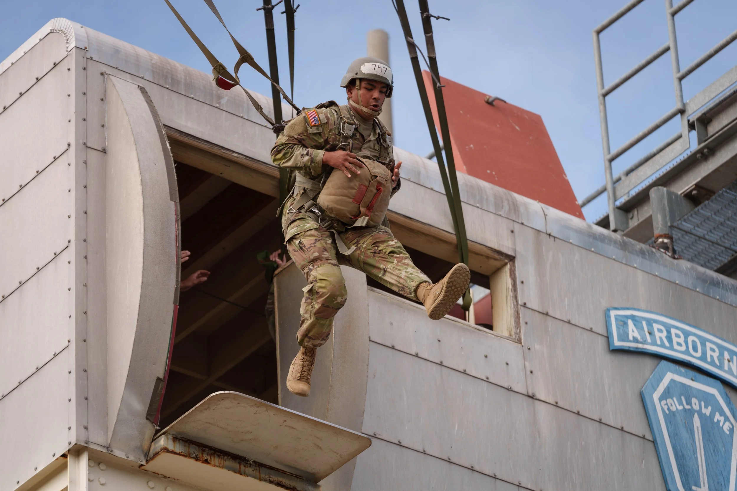 A soldier in camouflage uniform and helmet is jumping out of a helicopter, holding a backpack, during a military operation.