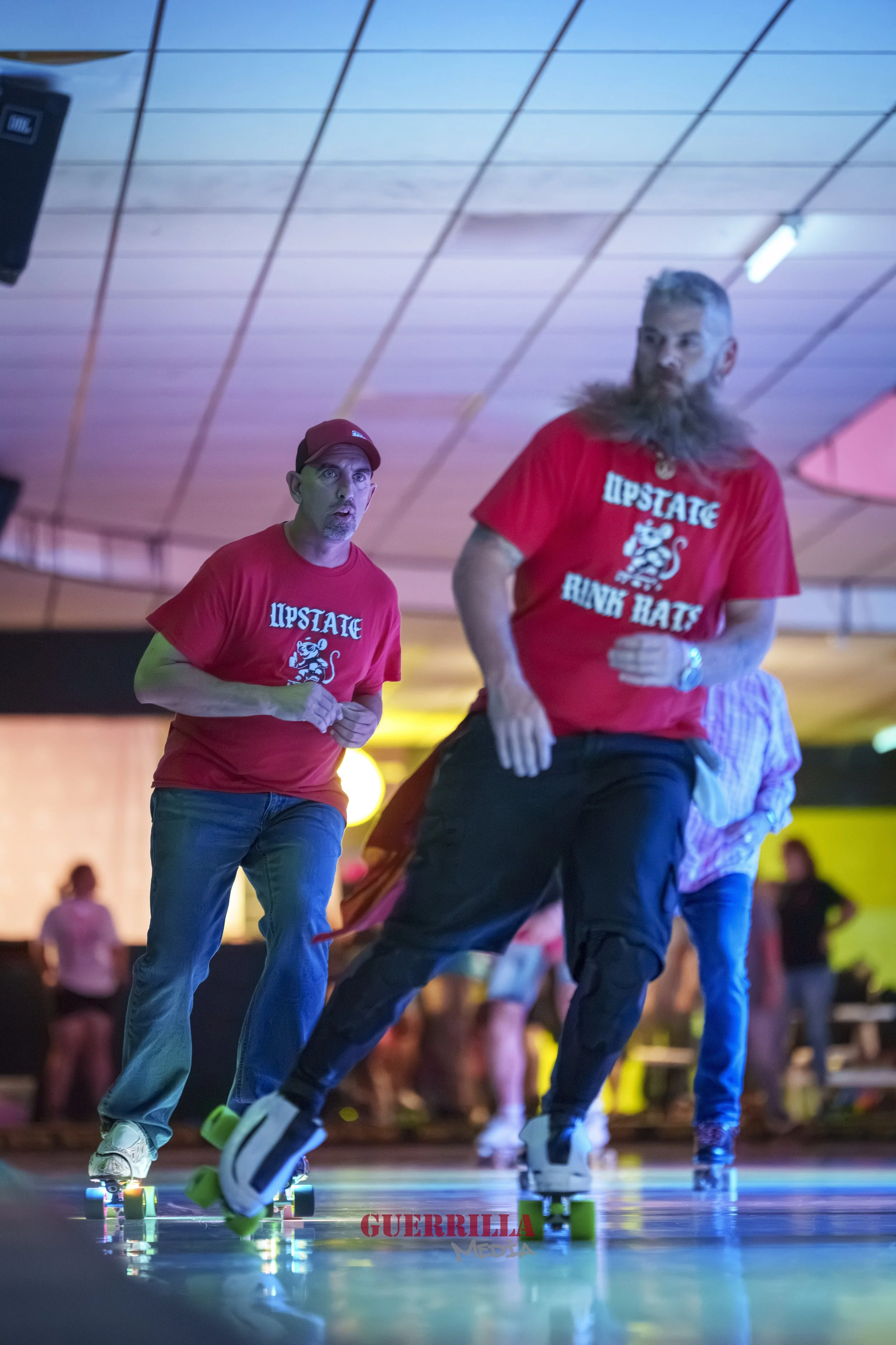 Two men skating indoors, both wearing red shirts with "UPSTATE RINK HATS" and a lion graphic, with one man wearing a red cap and the other with long hair and a beard, in a roller skating rink with colorful lighting and a few people in the background.