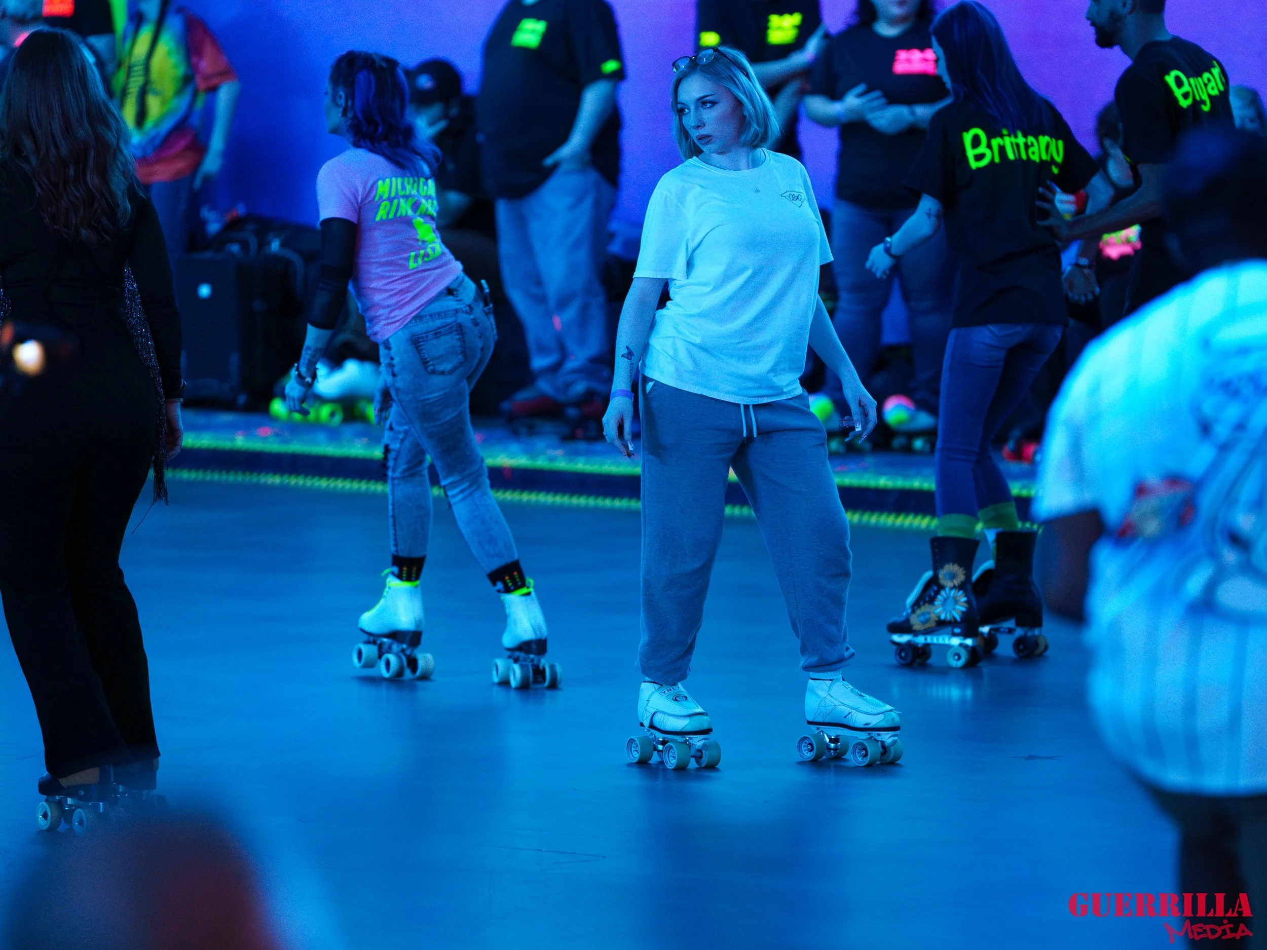 A group of people roller skating indoors under neon colored lighting, with some wearing shirts with neon names on the back, and others dressed casually. The setting appears to be a skate rink.