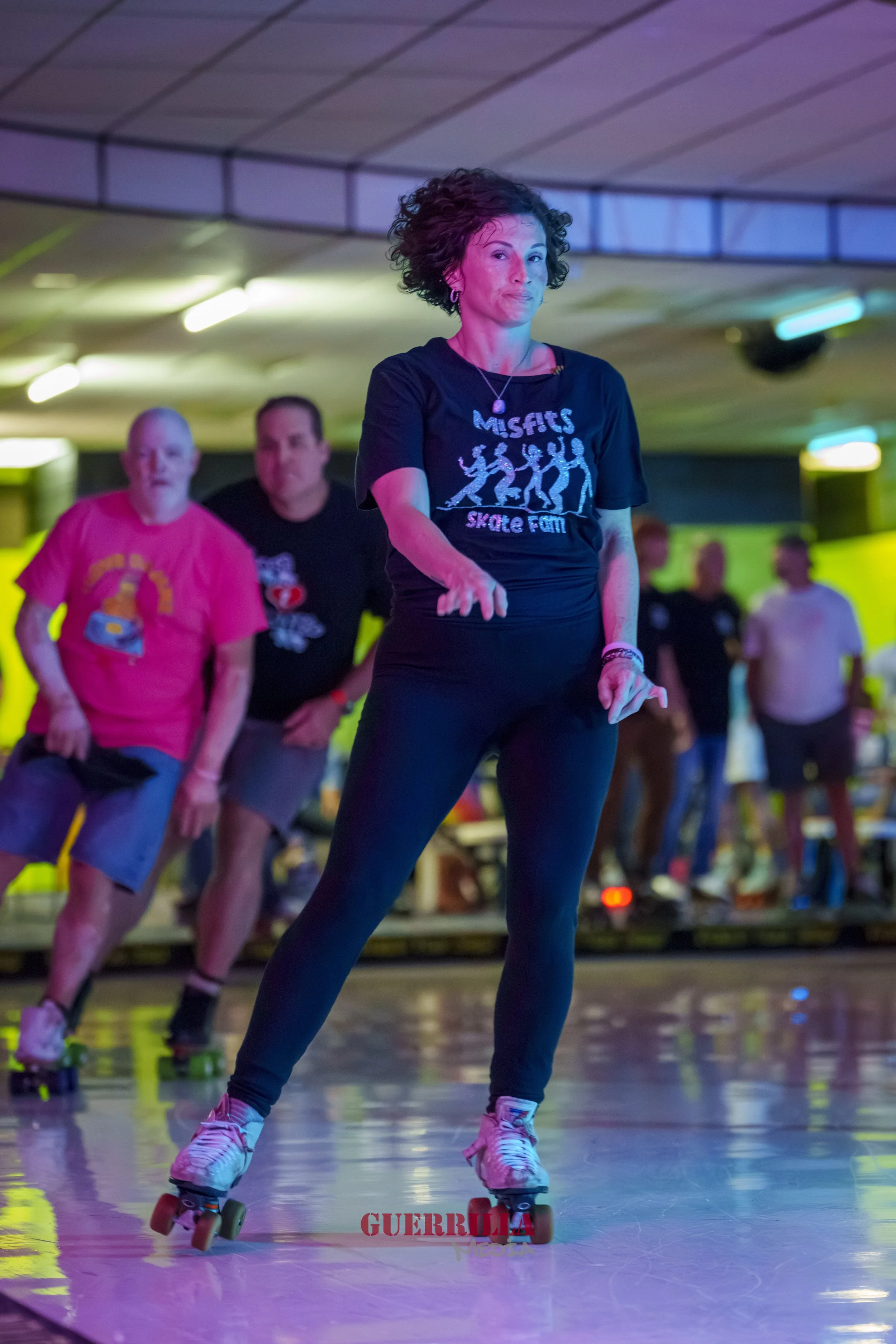 A woman roller skating indoors with a group of people in the background, some on roller skates.