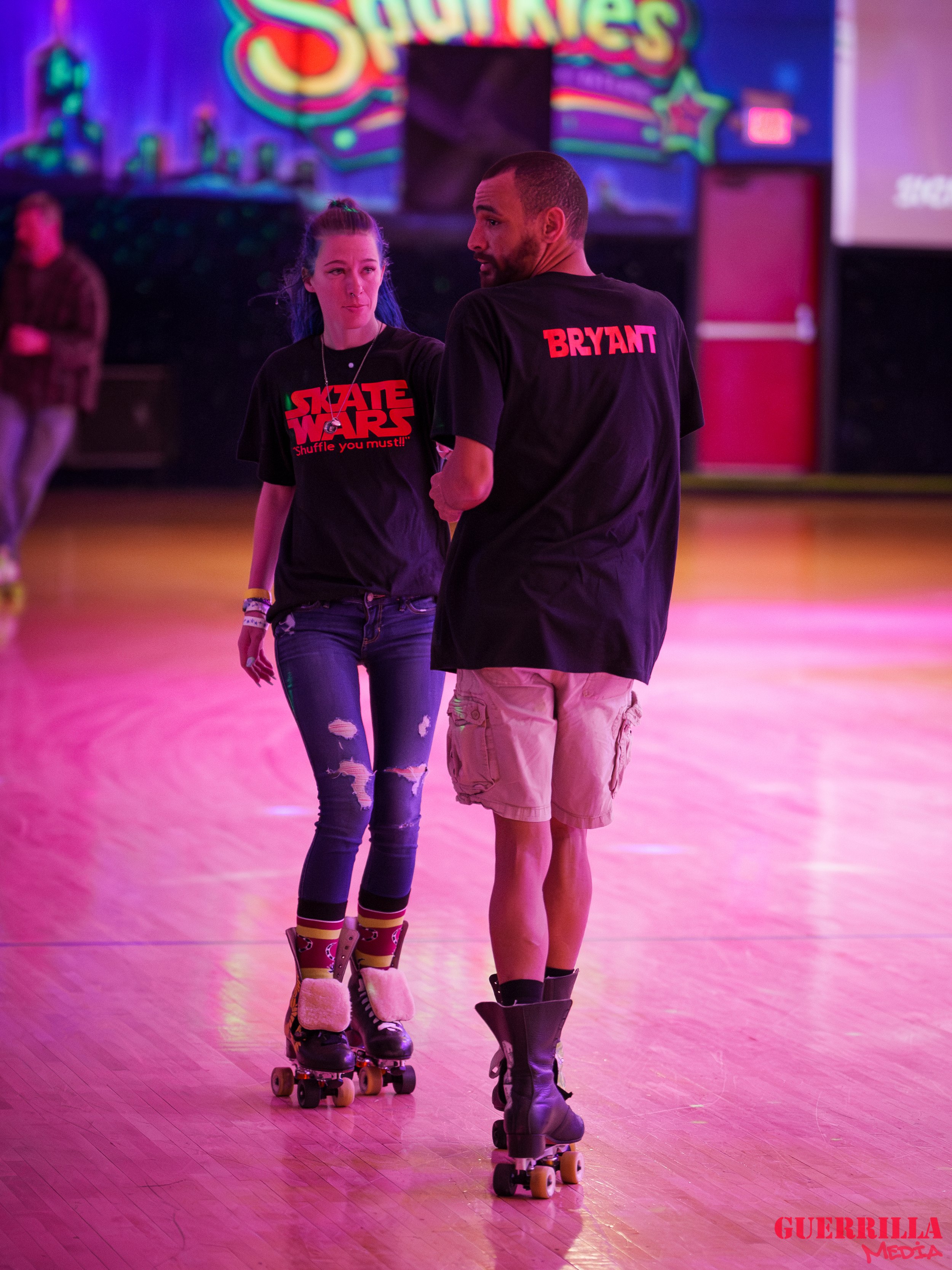 A young woman and a man roller skating together at an indoor skating rink with colorful neon lights and a 'Skate Wars' sign in the background. The woman has blue hair, is wearing a black 'Skate Wars' t-shirt, ripped jeans, and fuzzy roller skates. Th