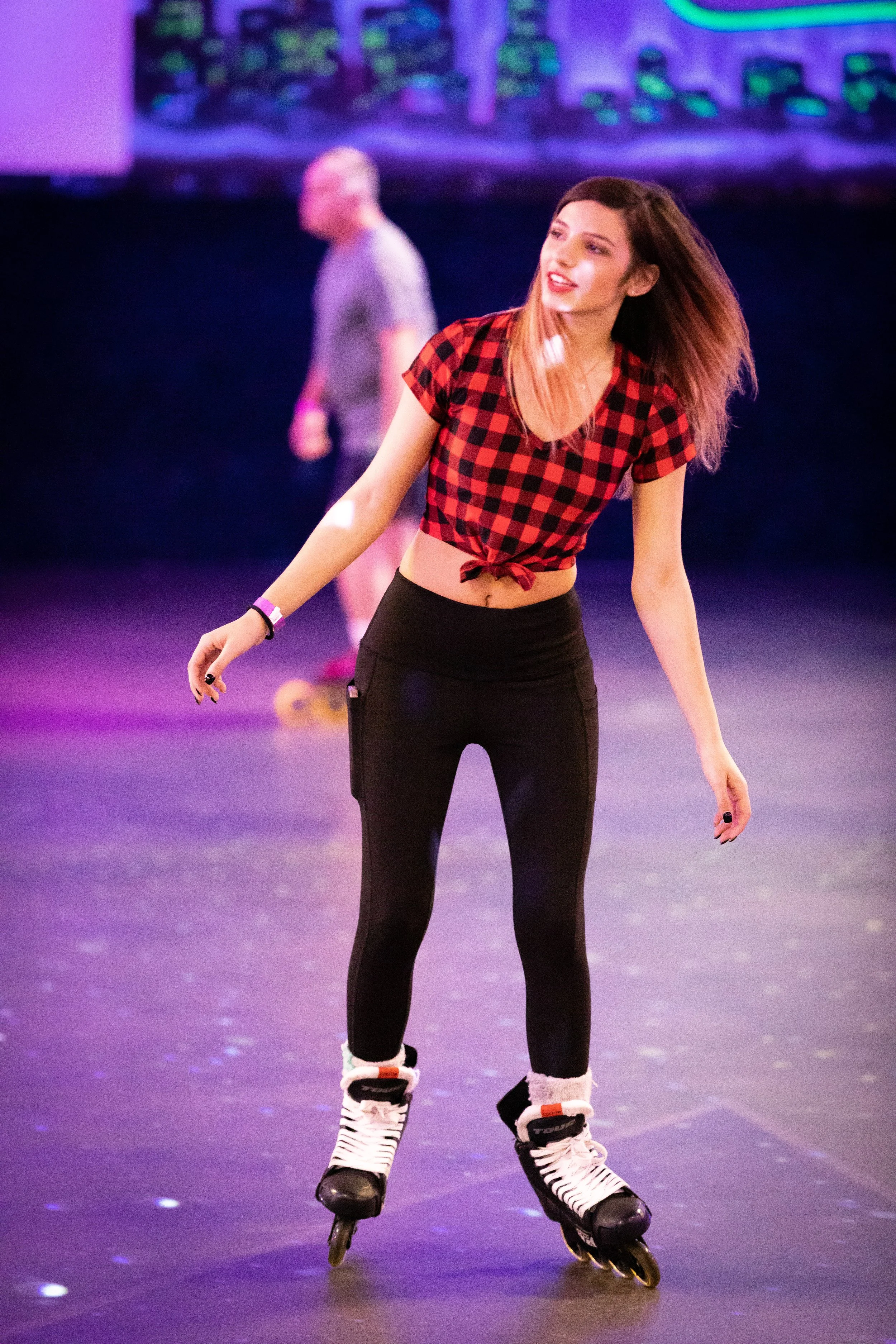 A young woman roller skating on an indoor rink, wearing a red and black checkered crop top and black pants, with colorful lighting and a cityscape backdrop.