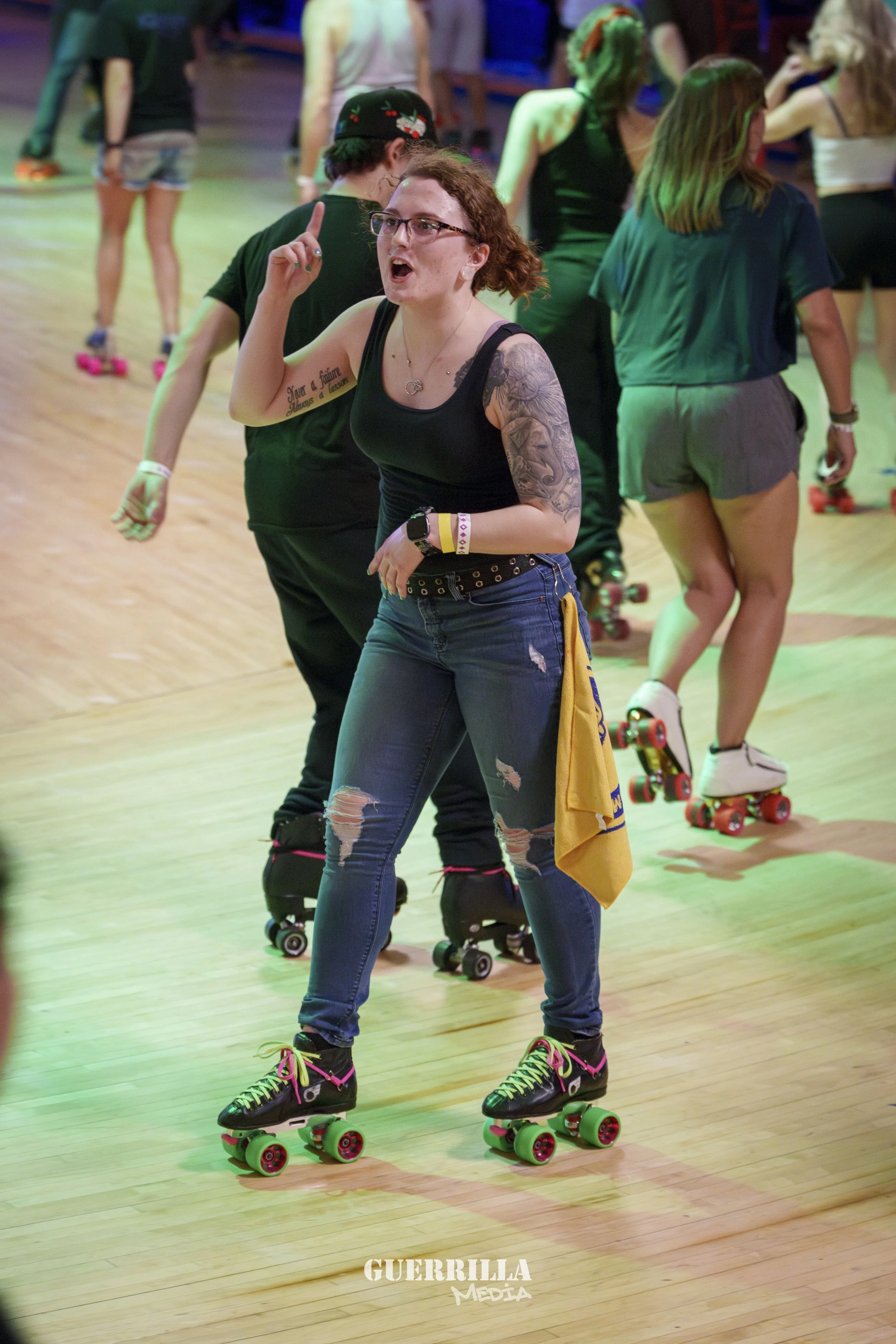 Group of people roller skating indoors, with a woman in the foreground talking or giving instructions, wearing black tank top, ripped jeans, and glasses.