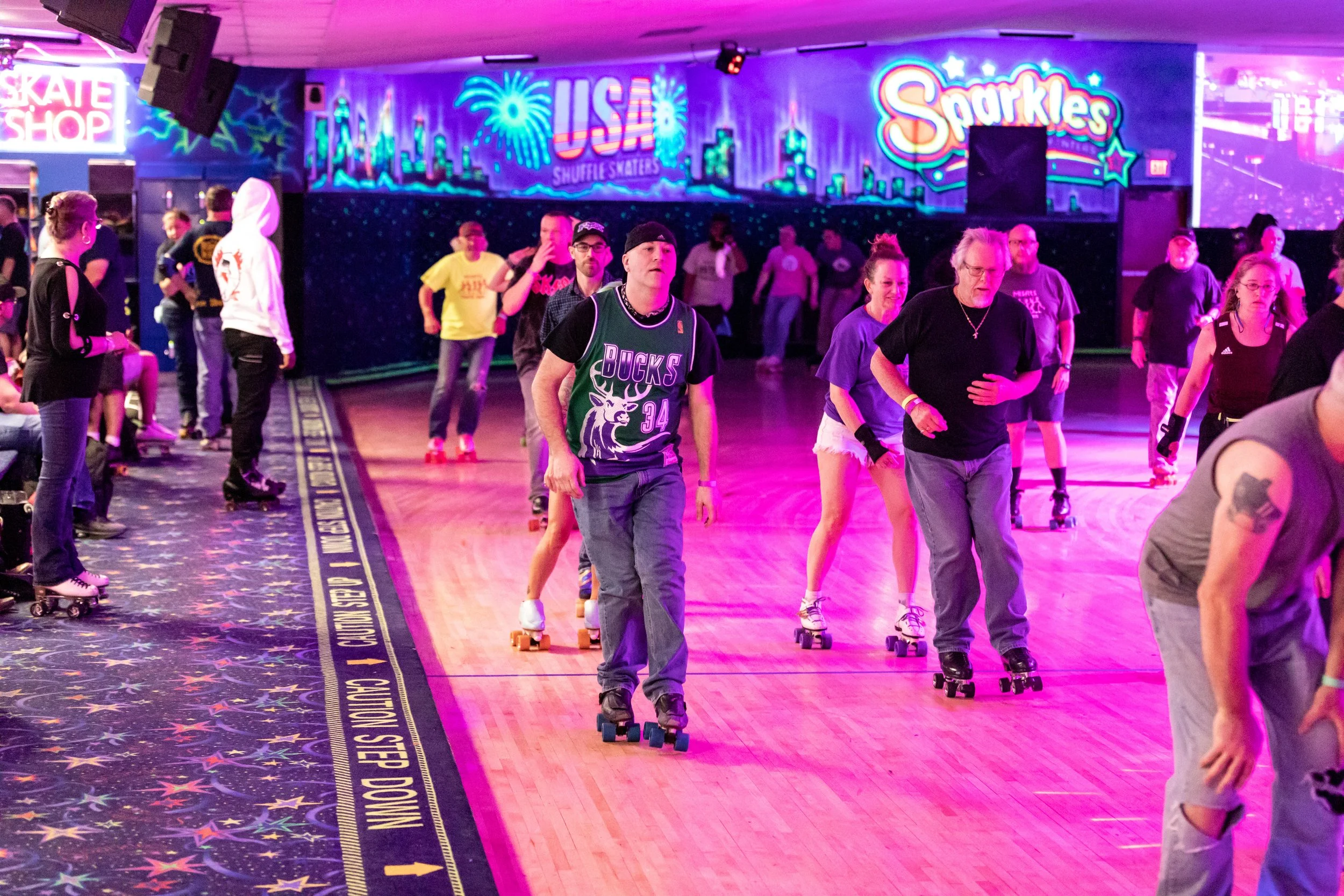 People roller skating indoors with colorful neon lights and advertisements for Sparkles and USA shuffle skaters in the background.