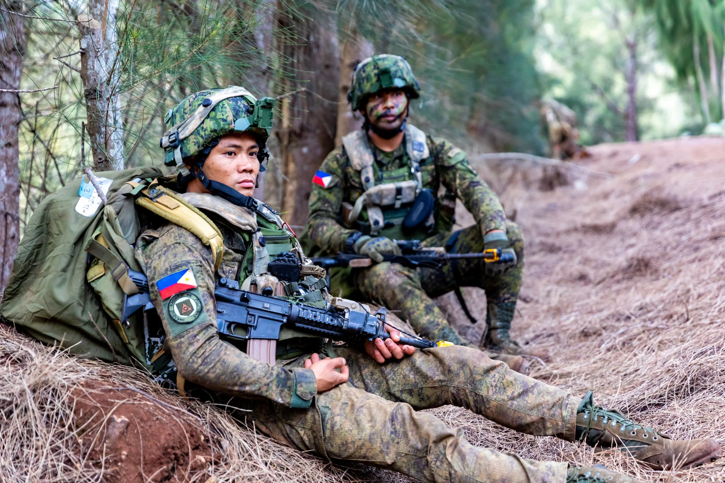 Two soldiers in camouflage uniforms with helmets and gear sitting on a forest floor among trees, one holding a rifle and the other holding a firearm, with patches indicating they are from the Philippines.