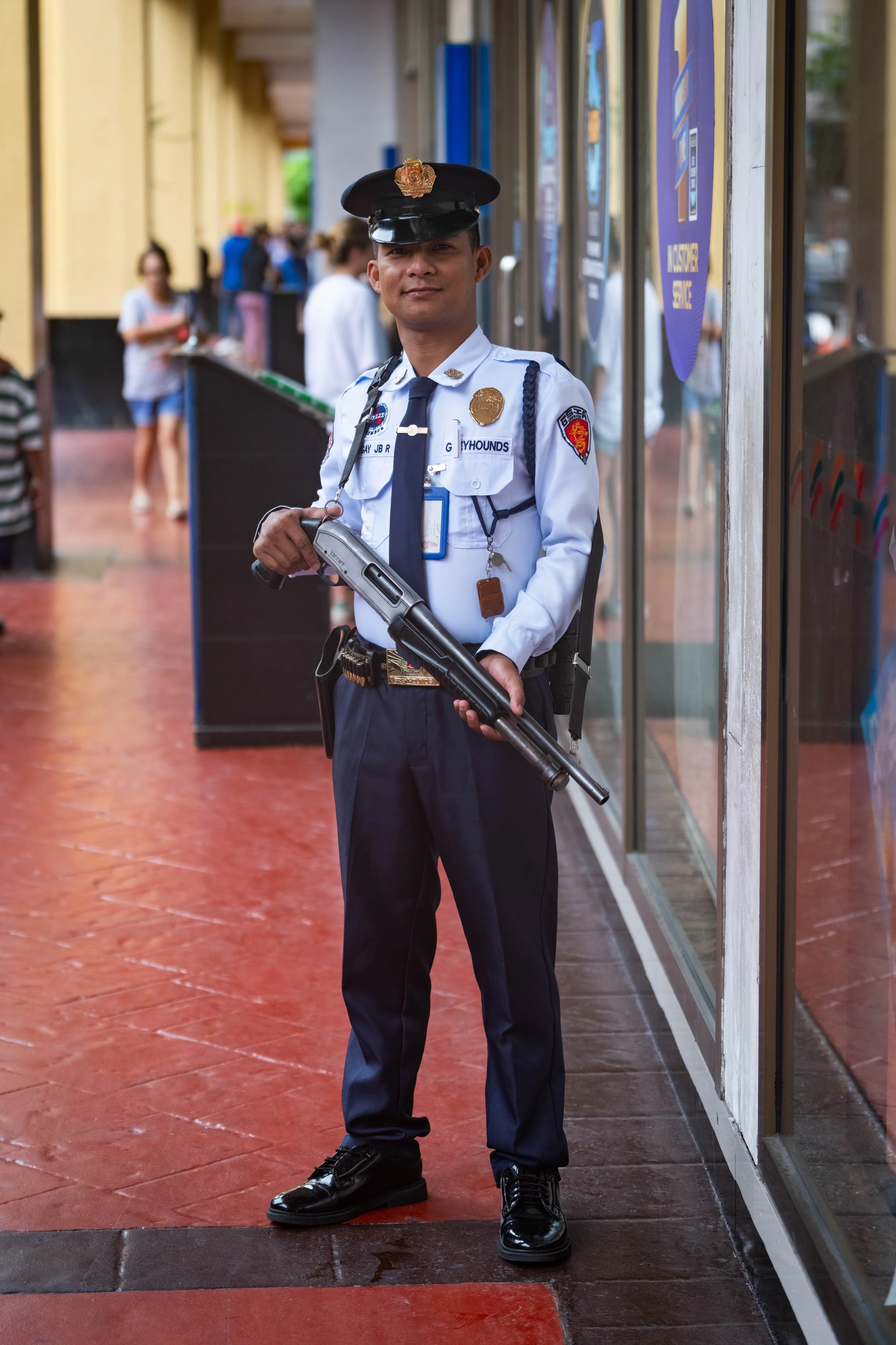 A security guard in uniform holding a shotgun, standing outside a building with people walking in the background.