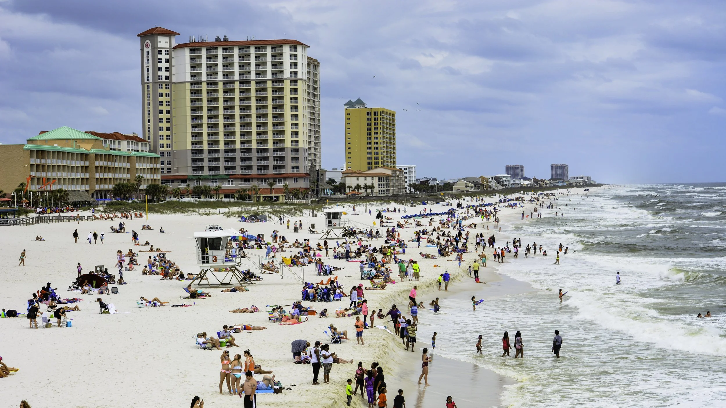 beach scene with many people sunbathing, swimming, and walking along the shoreline, high-rise buildings in the background, and a cloudy sky overhead