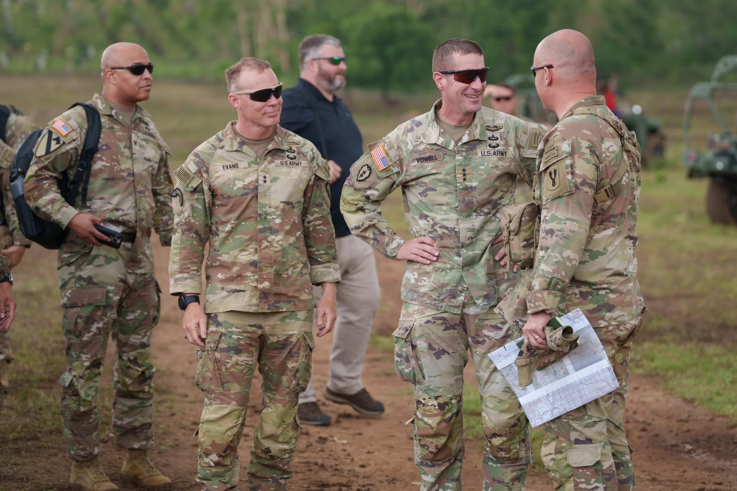 Group of U.S. Army soldiers in camouflage uniforms talking outdoors, some with sunglasses, with a man in civilian clothes in the background.