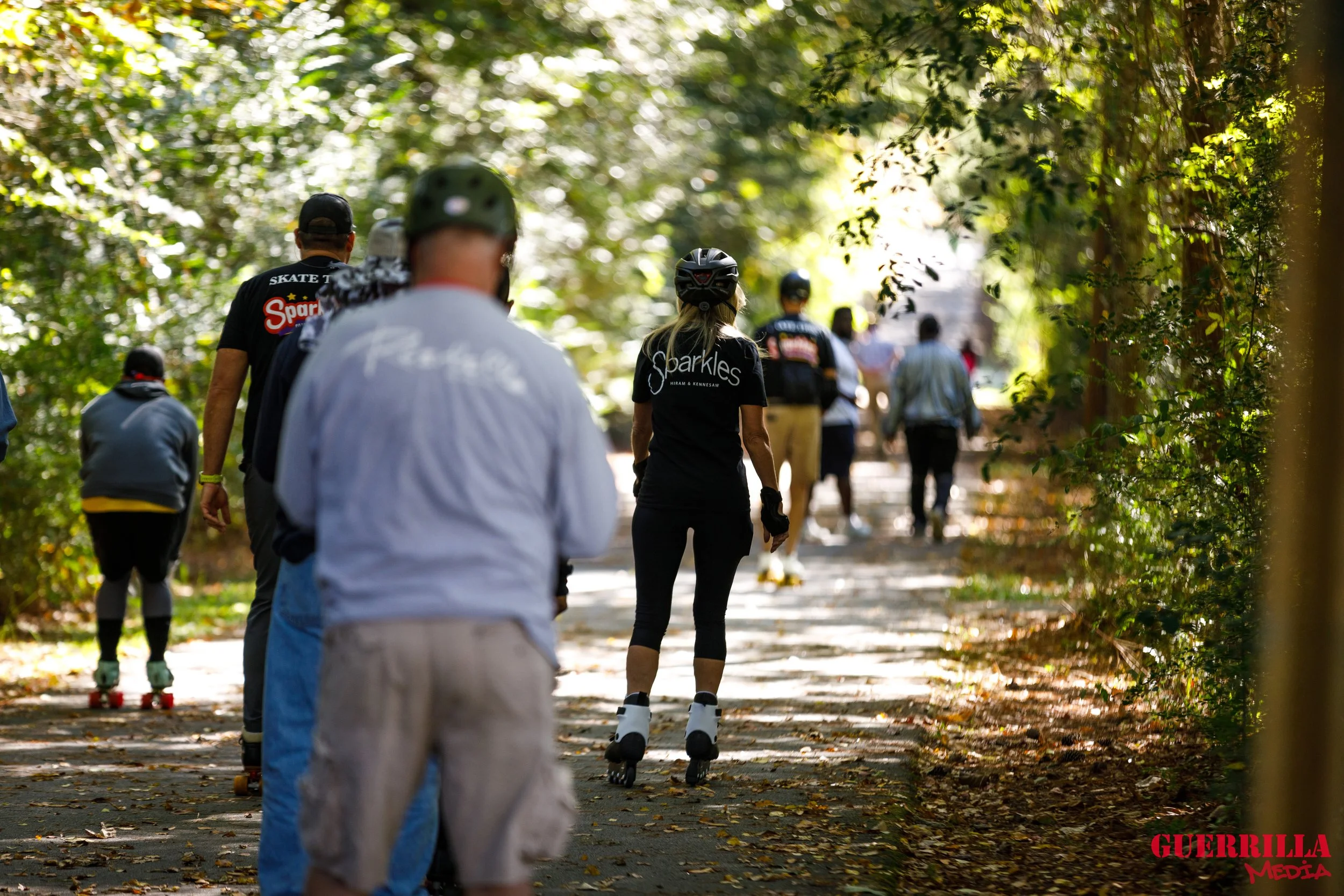 People rollerblading on a shaded trail through a wooded area.