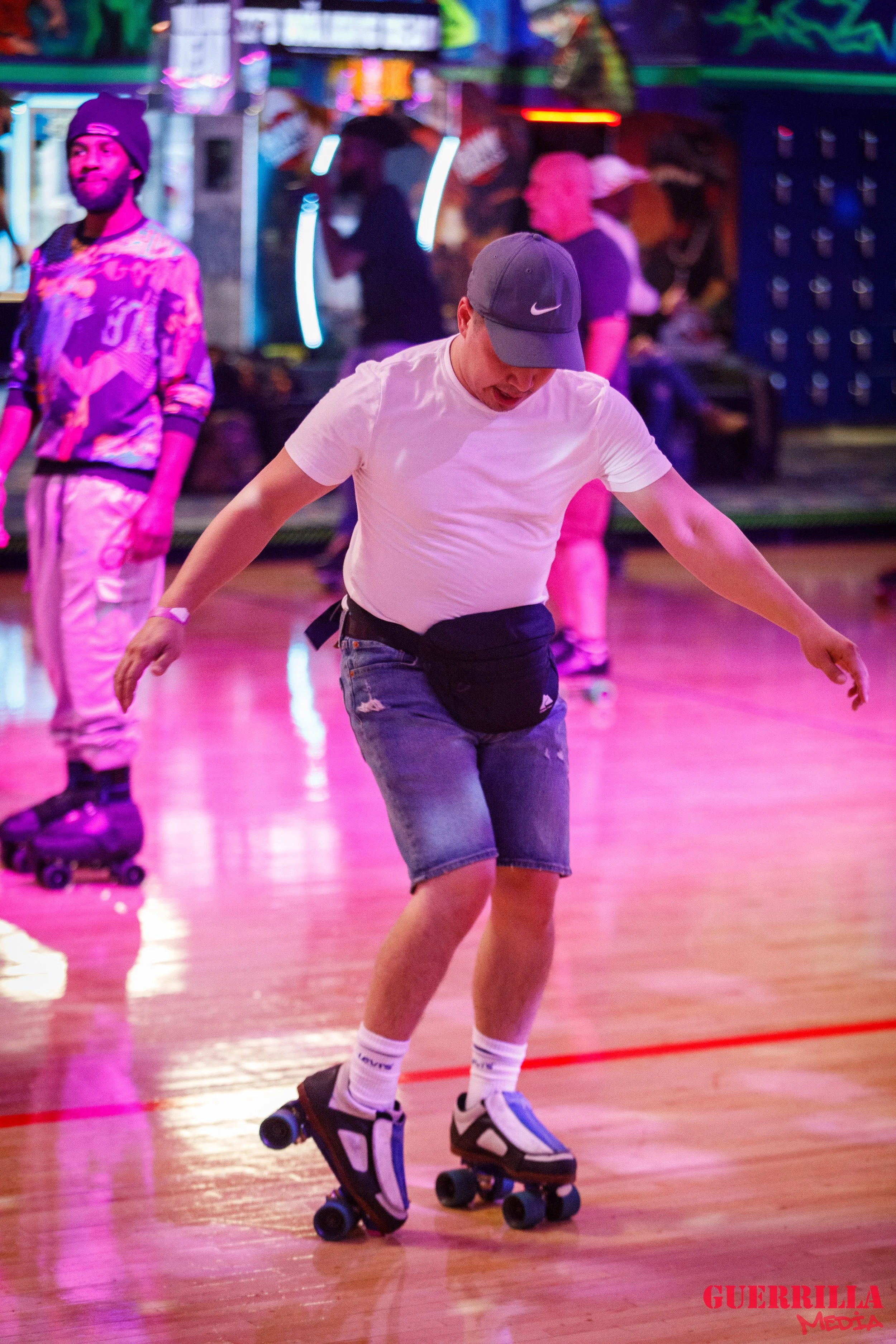 Person roller skating in a neon-lit indoor arena, wearing a white t-shirt, denim shorts, a dark cap, and a black waist pouch, with other skaters and colorful neon lights in the background.