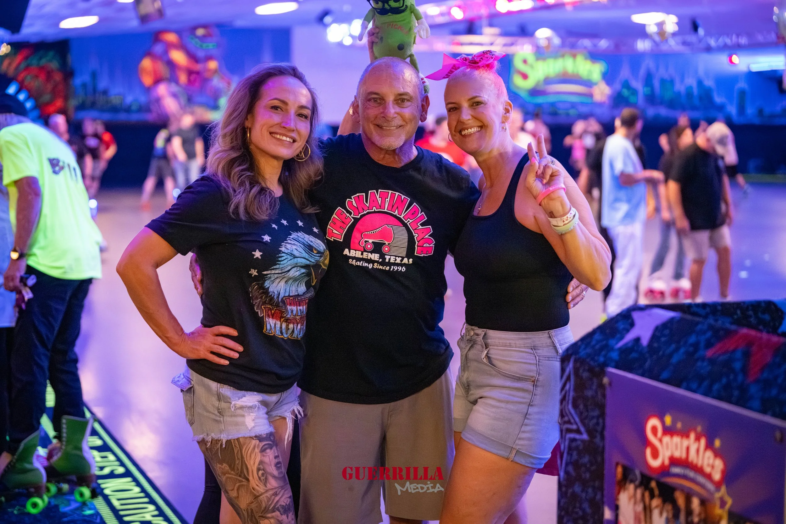 Three people smiling and posing together at an indoor roller skating rink, with other skaters in the background and a brightly lit sign that says 'Sprinkle.'