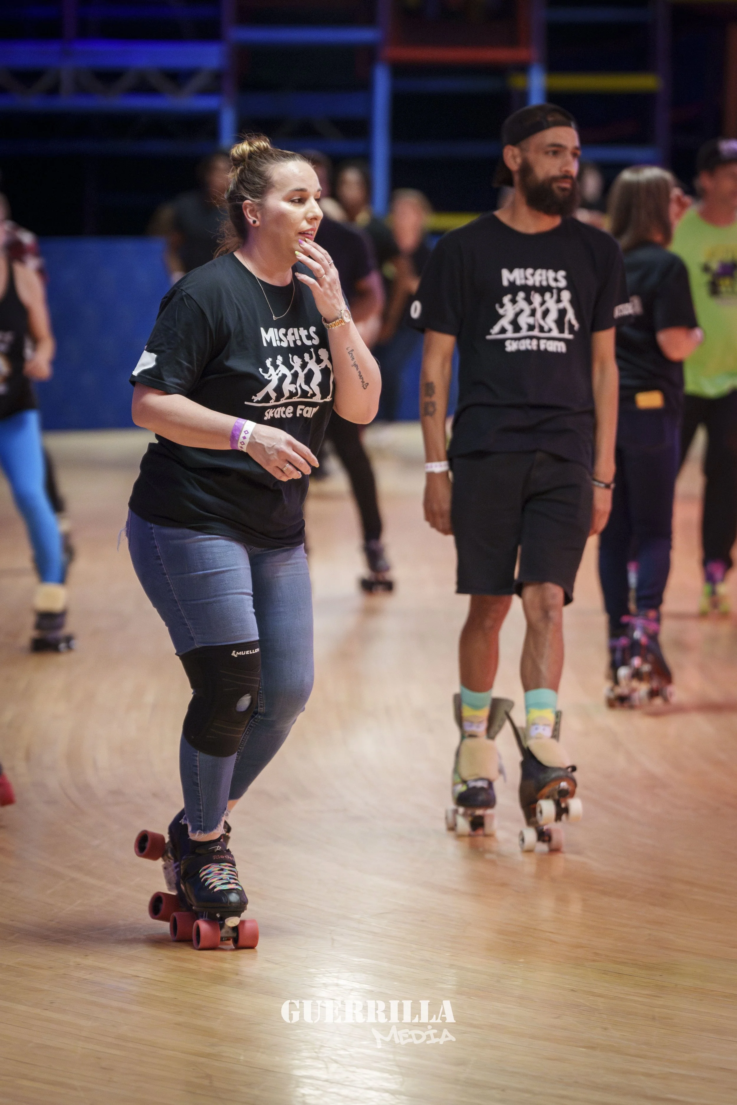 People roller skating indoors, wearing black 'Misfits Skate Farm' t-shirts, some with knee pads, in a skate rink with wooden flooring and colorful structures in the background.