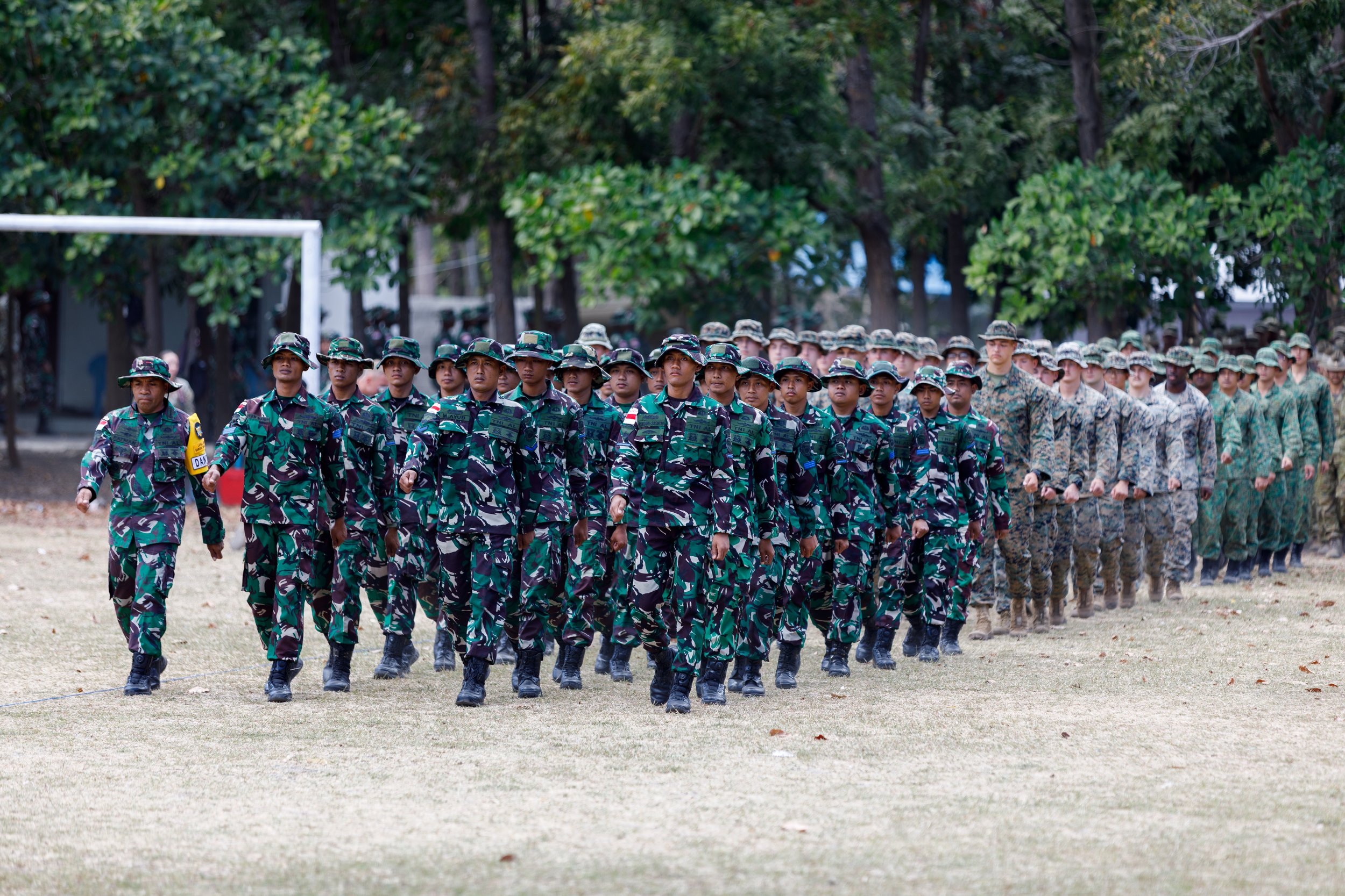 Group of soldiers in uniform marching in formation on a field with trees in the background.