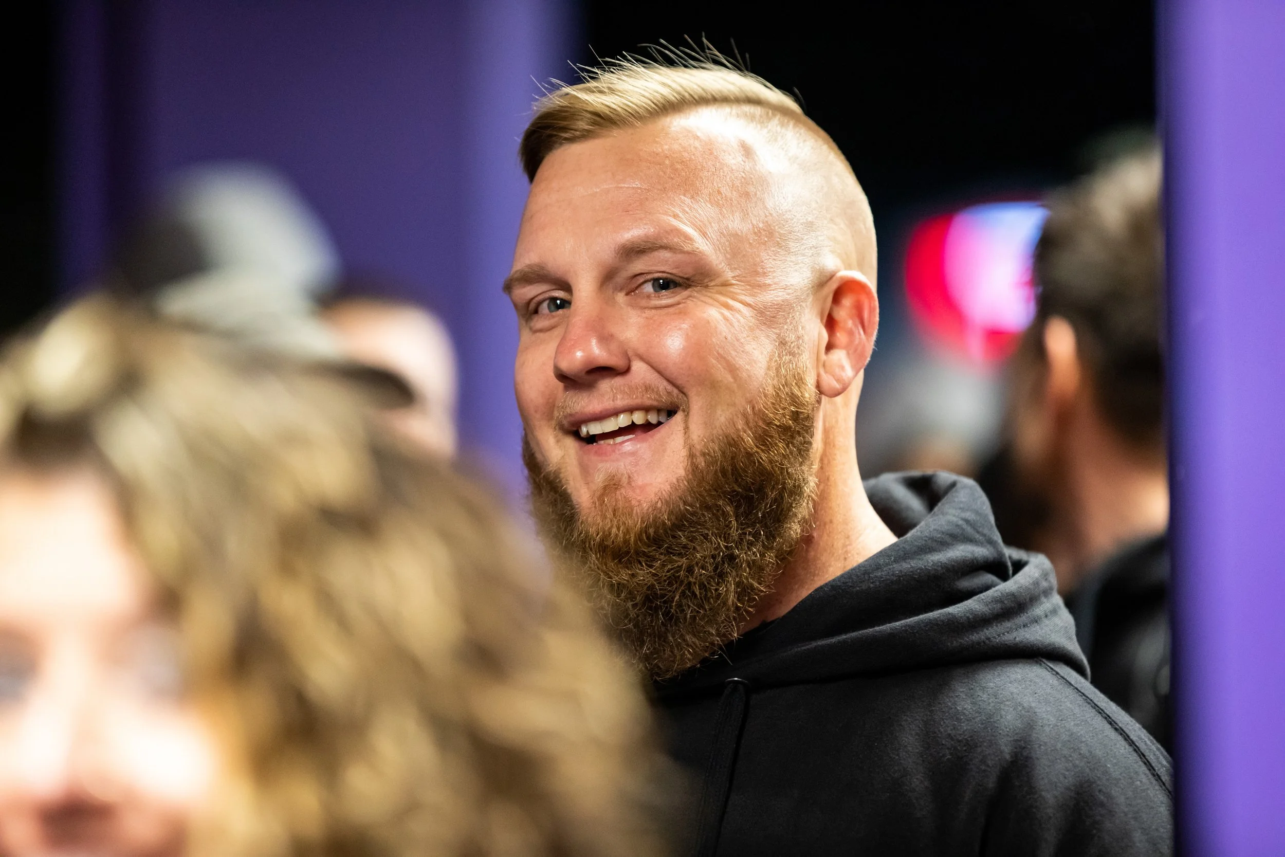 A smiling man with a beard and short blond hair, wearing a black hoodie, at a crowded indoor event.