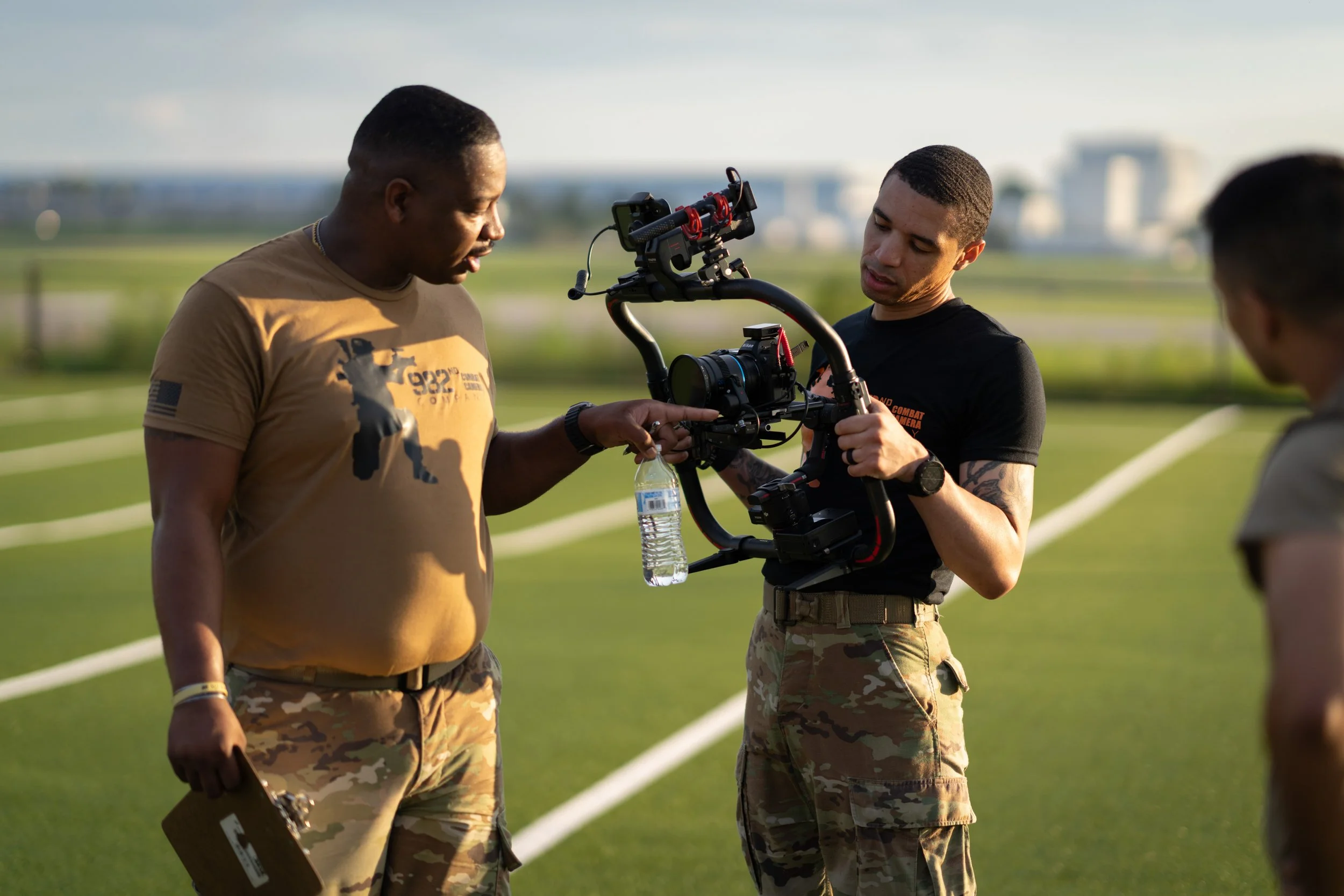 Three men in military cargo pants on a grassy field with white lines, one holding a water bottle, the other adjusting a camera stabilizer, while the third partially appears on the right, with a cityscape in the background.