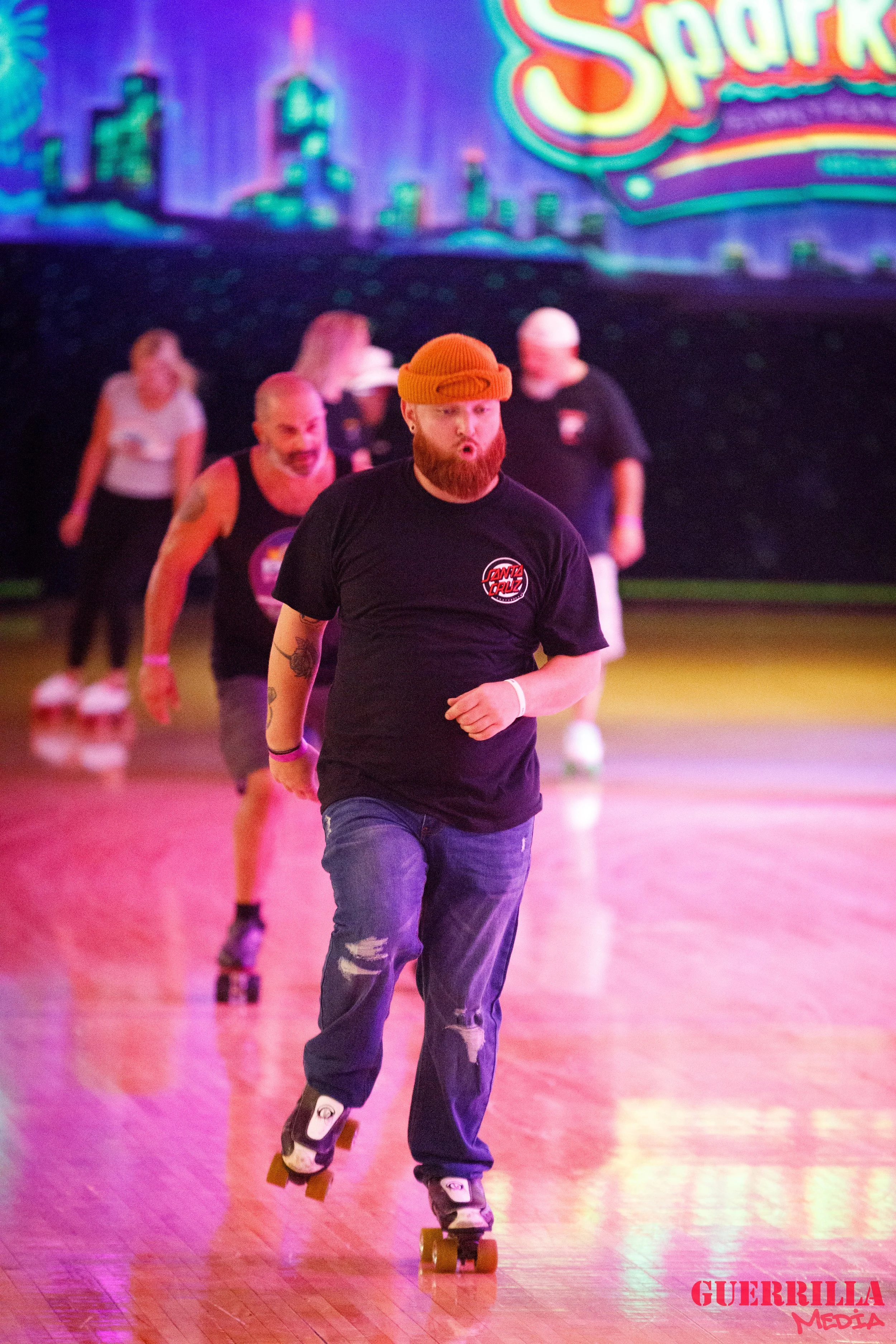 A man with a beard, wearing a red beanie and black t-shirt, roller skating indoors with a group of people in the background, with neon signs and city skyline graphics on a screen behind him.