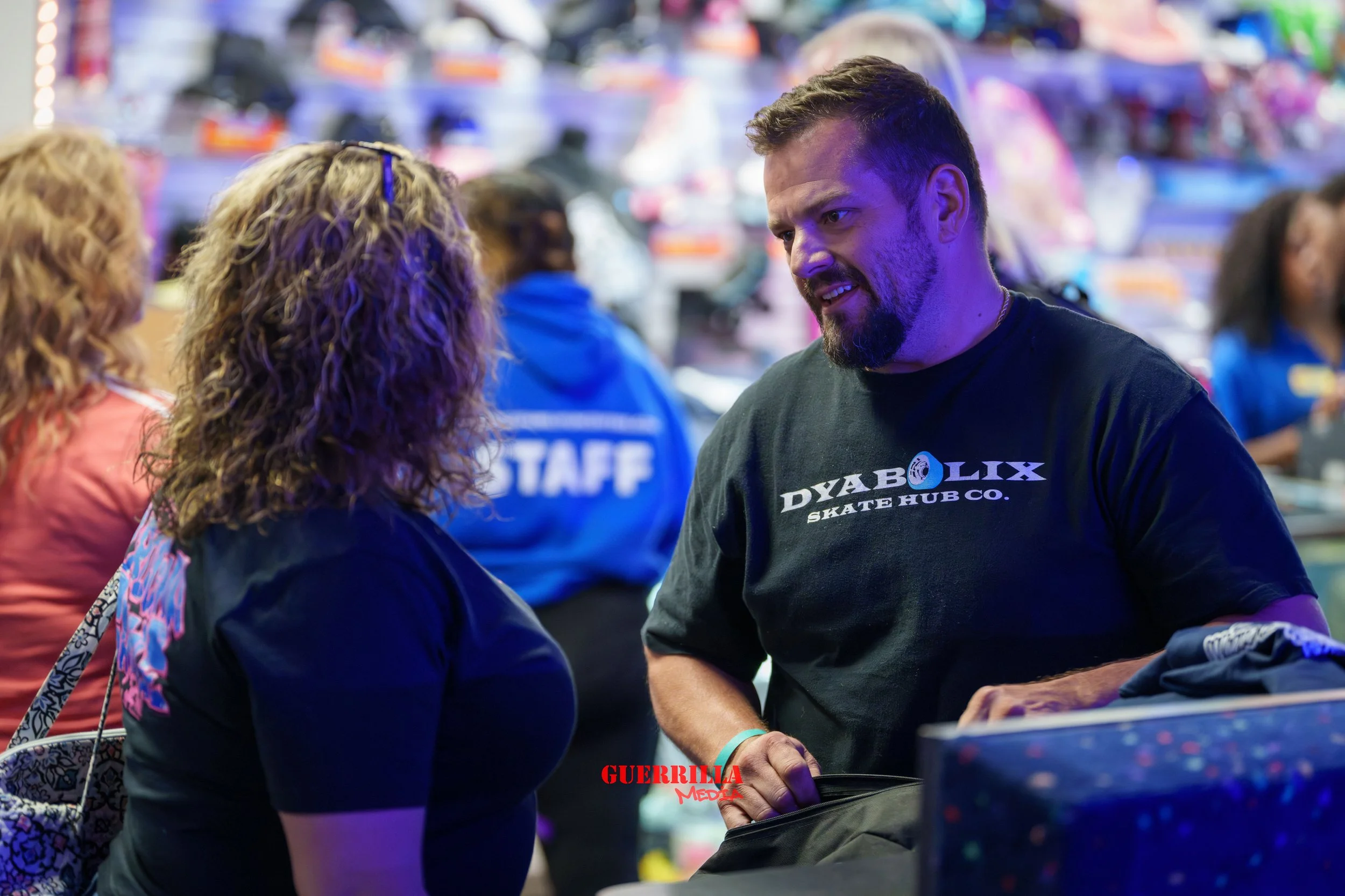 A man wearing a black T-shirt with the words "DYA BOLIX SKATE HUB CO." is talking to a woman with curly hair at a busy indoor skate shop or event. There are people and colorful items in the background, including a person wearing a blue staff shirt.